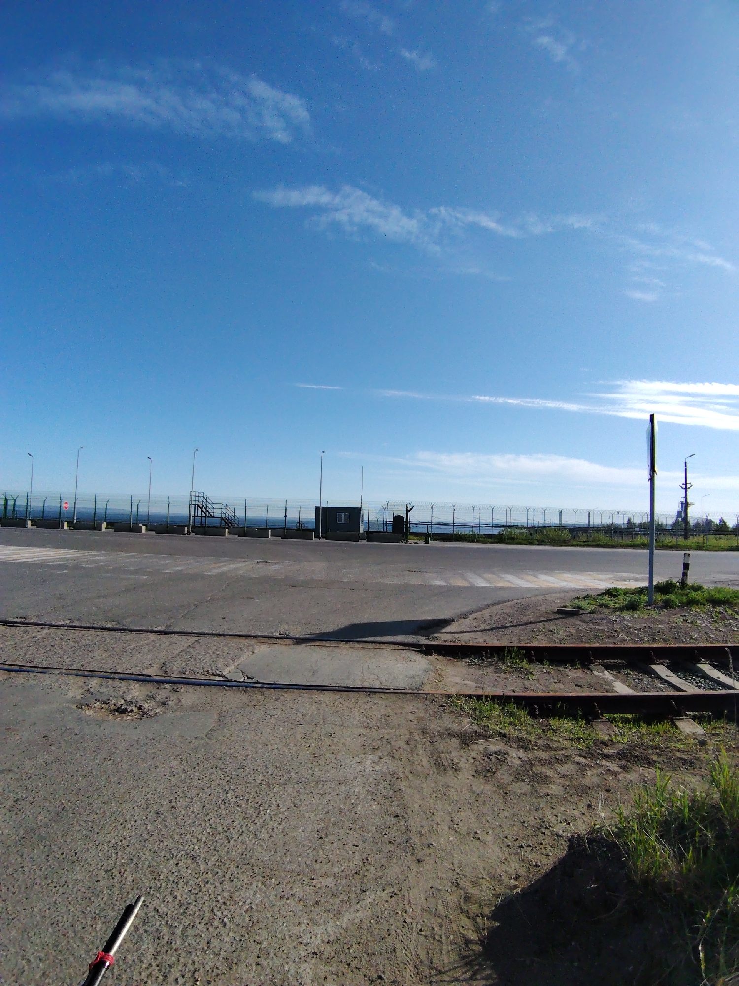 The scene is an outdoor view in Chaikovsky, Russia, showing a gray asphalt surface intersected by railway tracks with wooden ties and some weeds. In the mid-ground, there's a secured area with a fence topped with barbed wire, accessible via a metal staircase. Behind the fence is a body of water. Streetlights are evenly spaced along the fence. The sky is clear with sparse clouds. A thin cylindrical object with a red band is visible in the bottom-left foreground.