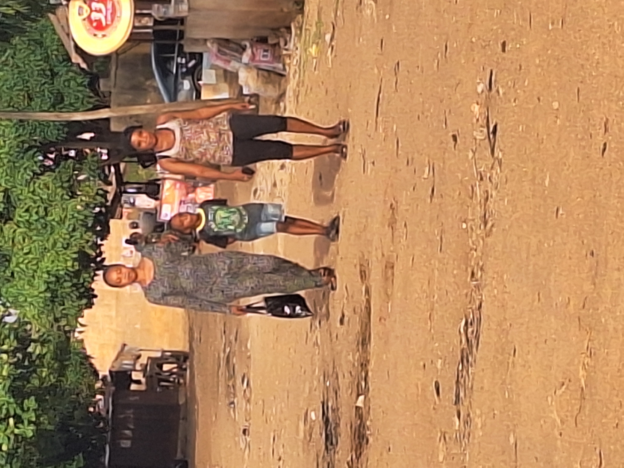 Three people are standing on a dirt road in Sabon Tasha, Nigeria. A woman is wearing a patterned dress and carrying a black bag. A child is wearing a t-shirt and denim shorts. Another woman is wearing a tank top and capri pants. A small store and dense foliage are visible in the background.