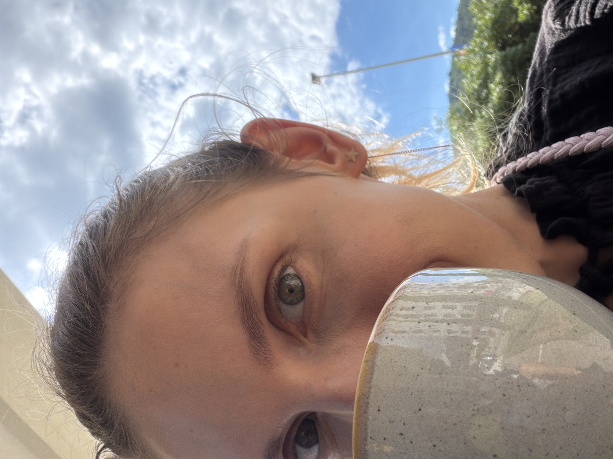 The image depicts a close-up, tilted view of a person's face, partially obscured by what appears to be a light grey ceramic mug. The person has light skin, fair hair, and hazel eyes. A small gold star-shaped earring is visible in the left ear. The person is wearing a black garment with pink braid detailing. The background shows a partly cloudy sky, and a utility pole is visible. Vegetation and a glimpse of buildings suggest a location within a populated area. Based on the presence of vegetation and buildings in the background, and the proximity to an urban center, this image may have been taken in or near Budva, Montenegro.