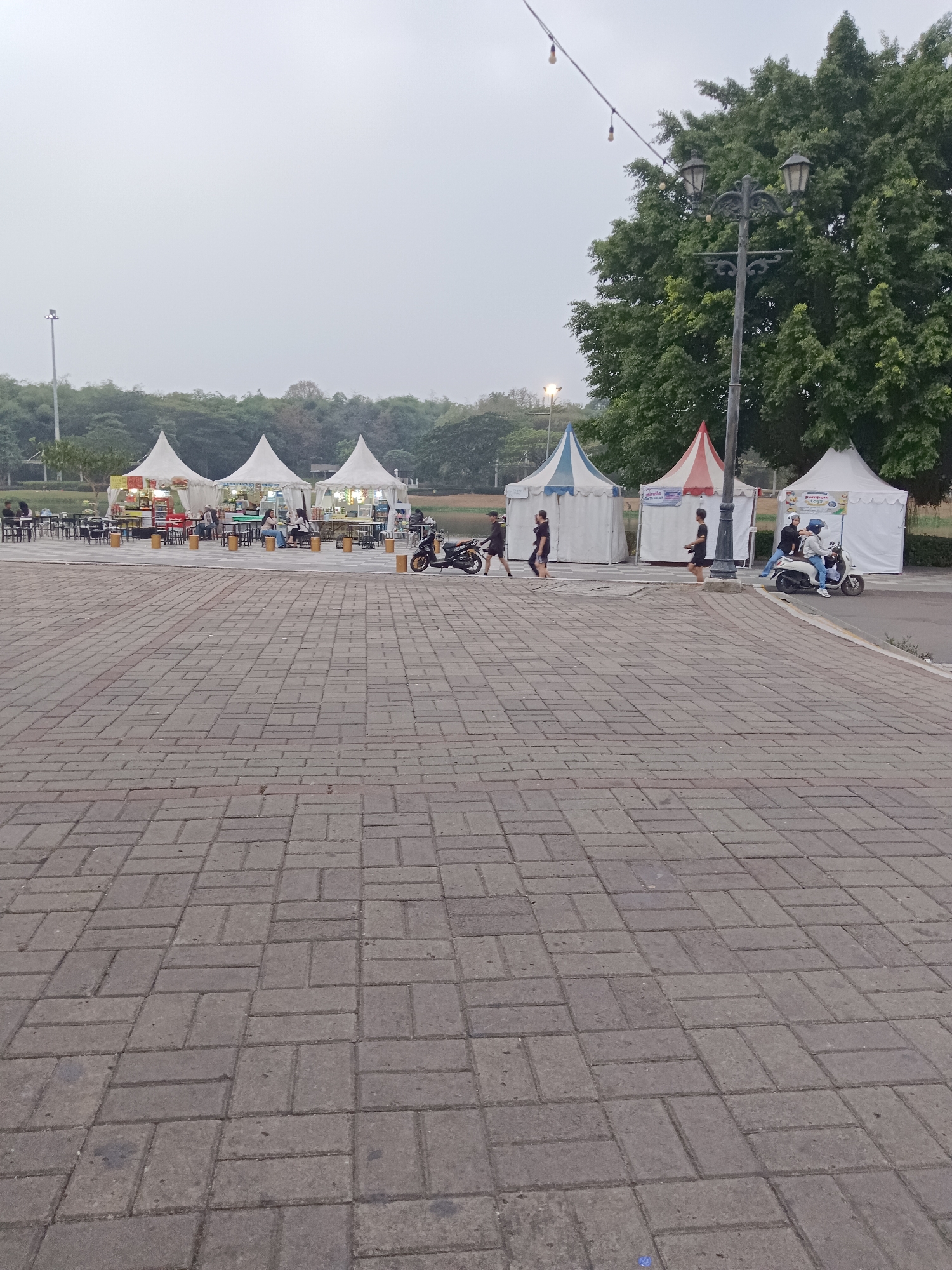 This image depicts an outdoor scene, likely a food court or market area, in the city of Jayamukti, Indonesia. The foreground is dominated by a wide expanse of grey interlocking paving stones. In the mid-ground, a row of white, conical-shaped tents are set up. Some tents have tables and chairs arranged in front of them, indicating dining or seating areas. Several people are visible walking or interacting within this area. One person is riding a white scooter, while another is on a black motorcycle. A lamppost with multiple lights stands prominently near the tents. In the background, lush green trees and foliage create a natural backdrop, suggesting the location might be near a park or natural landscape. The sky is overcast, contributing to a diffused light.