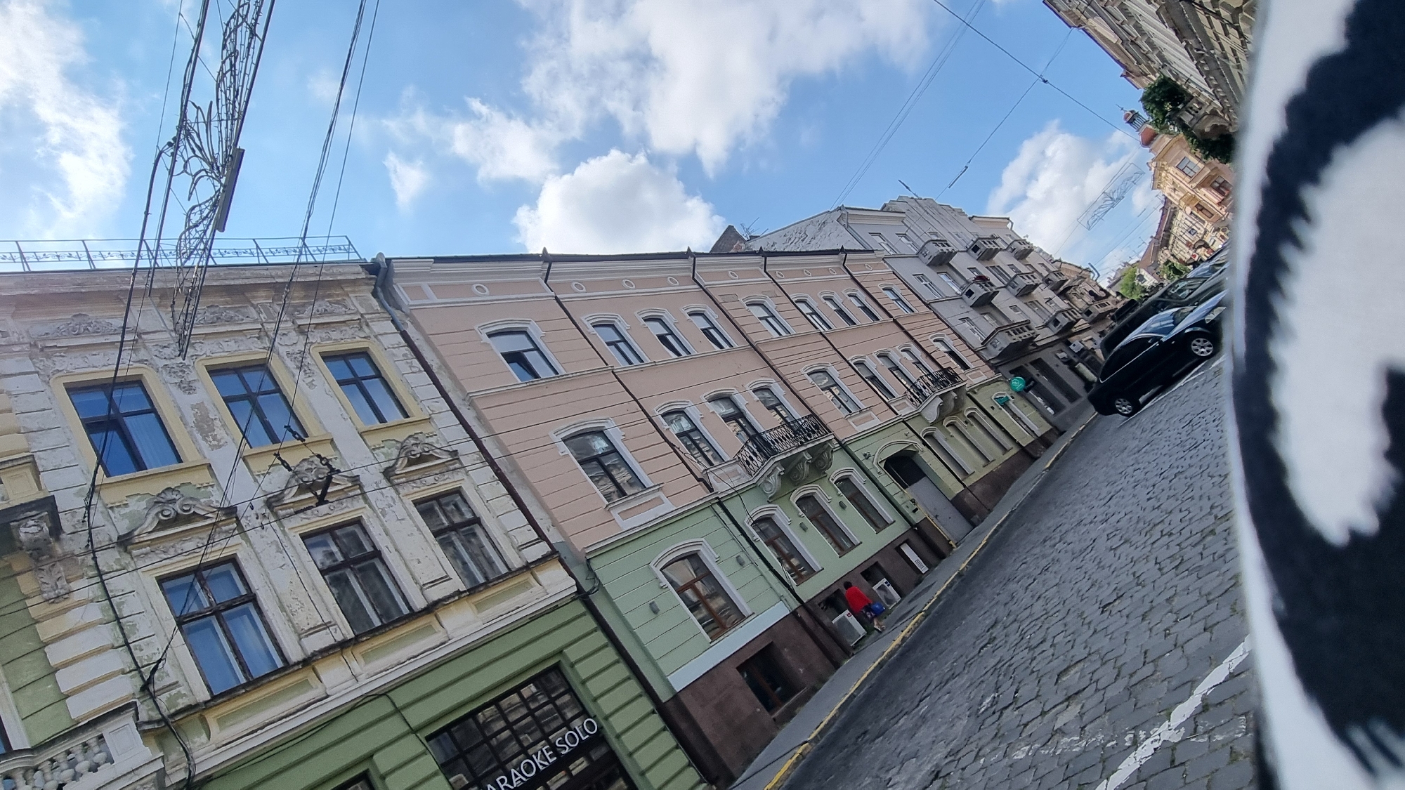 The image depicts a street scene in Chernivtsi, Ukraine, characterized by the angled perspective of old European architecture and a cobblestone street. The buildings lining the street exhibit a mix of faded pastel colors, including yellow, pink, and green, with ornate detailing around the windows and upper facades. Some buildings appear to be in a state of disrepair, with peeling paint and visible age.
In the foreground, a black car is parked on the right side of the street. The street itself is paved with dark cobblestones, and a white line marking a lane is visible on the left. Overhead, numerous electrical wires crisscross the sky, supported by poles, suggesting an urban setting. The sky is blue with scattered white clouds.
On the left side of the street, a green building with large windows is visible, with signage that reads "KARAOKE SOLO." Further down the street, on the left, a person in a red jacket is standing near the entrance of a building, next to some blue objects. The overall impression is that of a historic district with active street life.