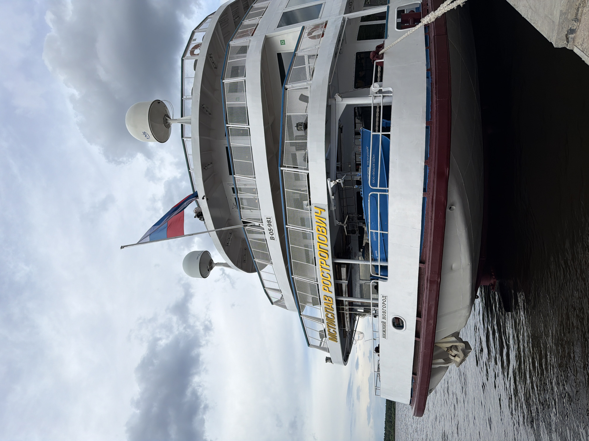 A white passenger ship, named "MSTISLAV ROSTROPOVICH" in yellow lettering, is docked at a pier under a cloudy sky. The ship has multiple decks with railings and windows. A Russian flag is visible at the top of the ship. Text on the ship reads "B-05-981" and "НИЖНИЙ НОВГОРОД". A blue life raft is visible on one of the decks. The water around the ship is dark and choppy. This image was taken in the city of Perm, Russia.