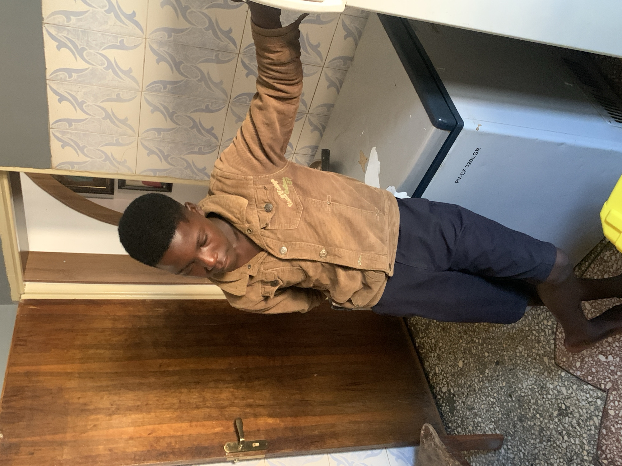 In a room with tiled walls and a wooden door, a young Black male stands near a freezer. He is wearing a brown corduroy jacket over a blue t-shirt and navy shorts. The freezer, labeled "PV-CF 320LGR," is light gray with a black top edge. The floor is a mosaic of stone and terrazzo. The environment suggests a domestic setting within a city, potentially Jos, Nigeria, given the cultural context.