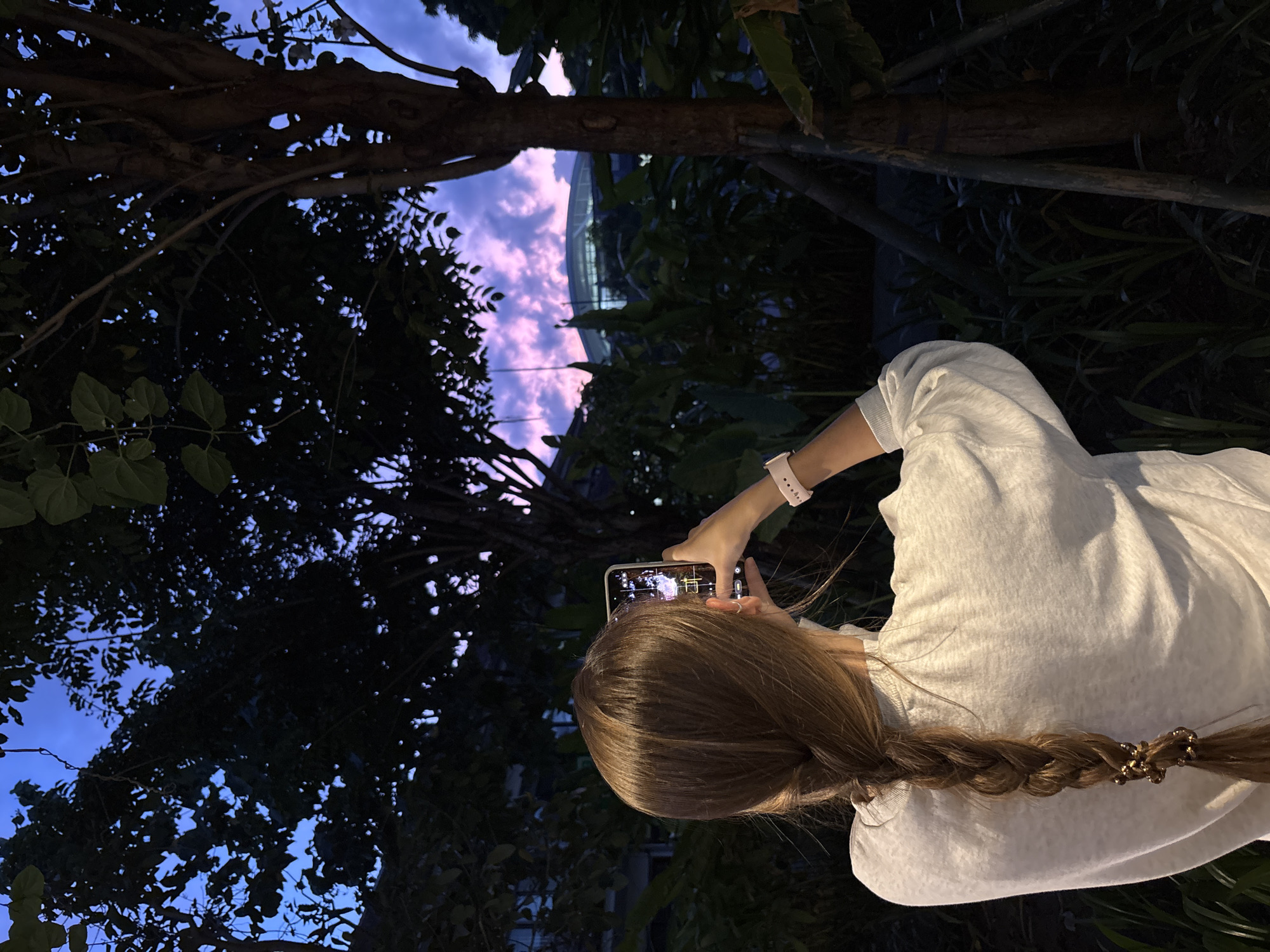 A woman with blonde hair in a braid, wearing a white shirt, is holding a smartphone up, taking a picture of the sky at twilight. The sky visible through the tree branches is a mix of blues and pinks. Behind the trees and sky, a large, curved building is partially visible. The photograph was taken in Kuta, Indonesia.