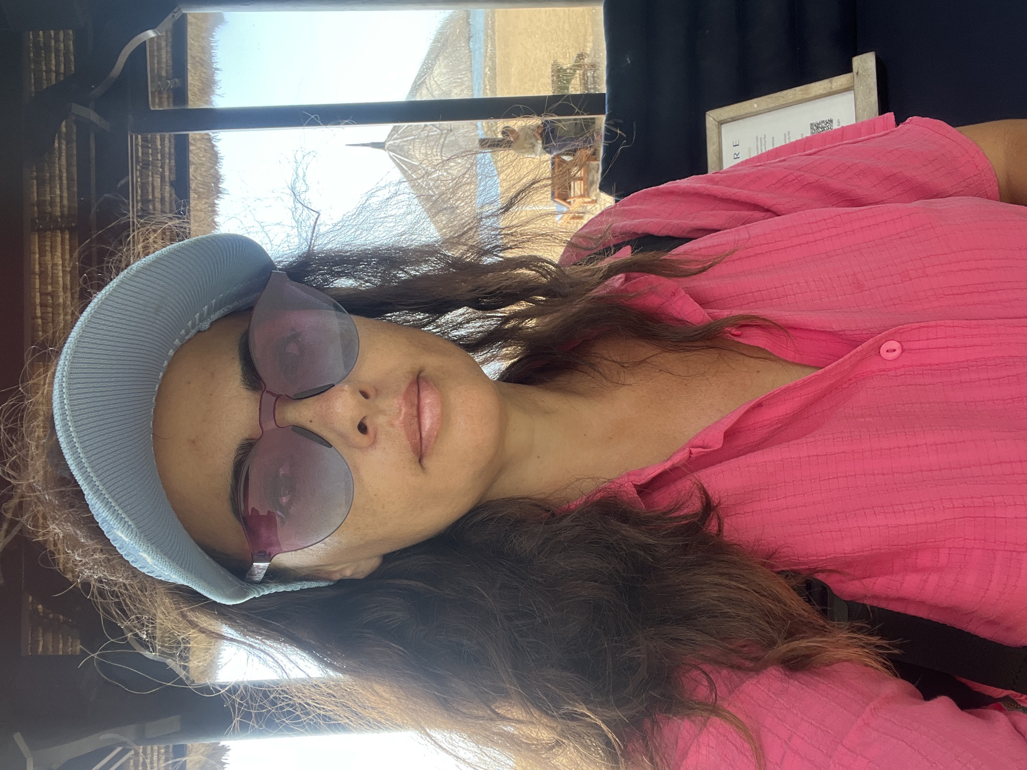 A woman is taking a selfie in Kuta Selatan, Indonesia, likely at a resort or hotel. She is wearing a pink button-up shirt and a light blue visor. Behind her, through a window or open doorway, a beach scene is visible with sand, lounge chairs, thatched umbrellas, and the ocean under a clear sky. The woman has long, dark, wavy hair and is wearing oversized pink-tinted sunglasses. The interior where she is standing appears to have a thatched ceiling.