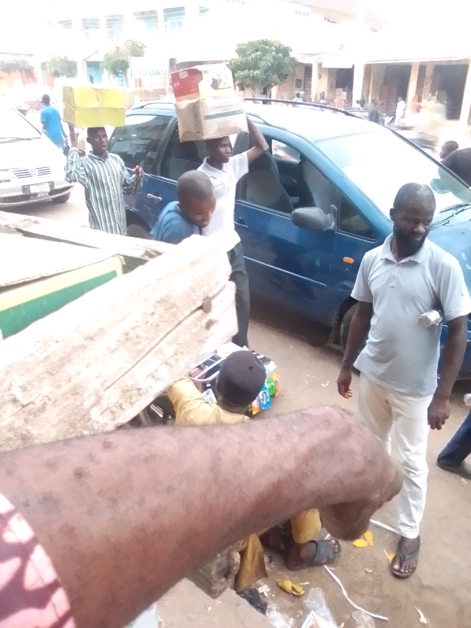 In Arawa, Nigeria, a man in a light blue polo shirt and white pants stands to the right, facing left, with his right hand holding a metal cylinder to his hip. Behind him, a blue minivan is parked. A man wearing a striped long-sleeved shirt and carrying a yellow box on his head walks towards the minivan from the left, his body angled towards the vehicle. In front of the minivan, a man in a blue shirt looks down and to the right. A large, wooden structure, possibly a stall or cart, is in the foreground on the left. Several people are visible in the background, and boxes are stacked on top of the minivan. The ground appears to be dirt or unpaved road.