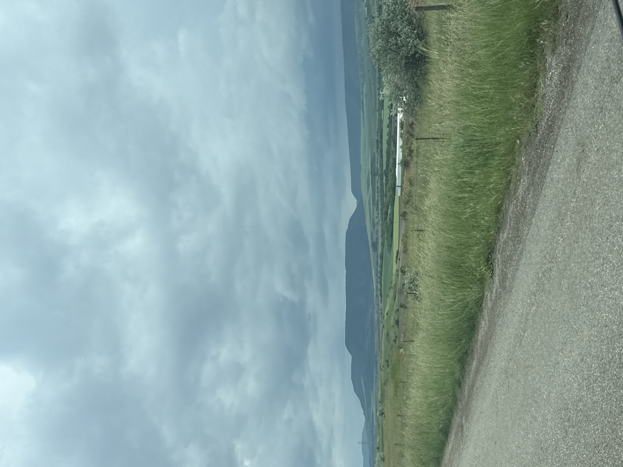 The image presents a landscape view. The scene is dominated by a cloudy sky that occupies a significant portion of the frame. Below the sky, there are mountains and flat land. Along the right edge, there's a road, followed by tall grass. A fence is visible, running parallel to the road and grass. There are trees and greenery in the distance, adding depth to the scene. The image does not provide enough context to definitively identify the exact location in the city of Kaspichan, Bulgaria, or any specific objects or actions.
