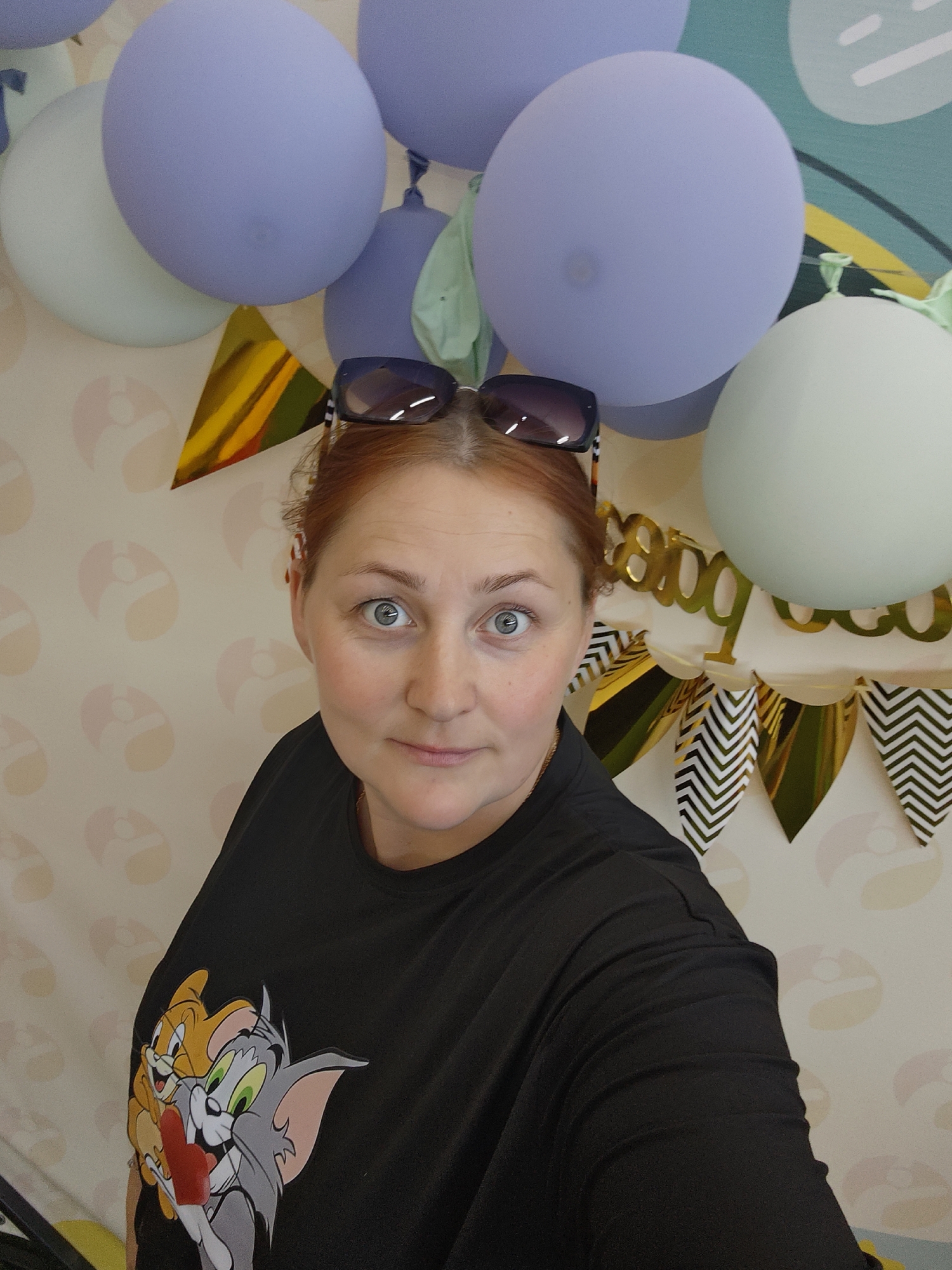 This media file is a portrait of a woman with red hair, wearing sunglasses on her head and a black t-shirt with an image of Tom and Jerry. She is looking directly at the camera with wide, surprised eyes and a slight smile. The background appears to be decorated for a celebration, with several balloons in shades of blue and green. There is also a gold banner with black and white chevron patterned pennants, and a gold banner that appears to spell "BOO". The wallpaper in the background has a pattern of stylized figures. The lighting suggests it could be daytime. The woman's expression and the festive decorations indicate a celebratory event, possibly a birthday. The location context provided is Chaikovsky, Russia.