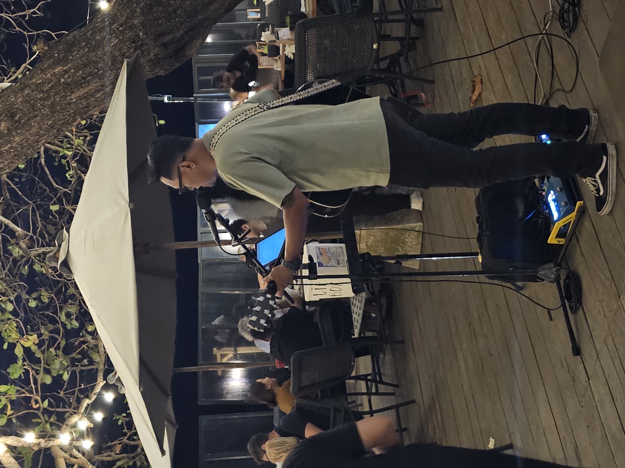 A man plays an acoustic guitar and sings into a microphone at an outdoor venue, possibly a restaurant or bar, in Denpasar, Indonesia. He is wearing a light green t-shirt, dark pants, and Vans sneakers. Behind him, diners are seated at tables under a large umbrella. Strings of fairy lights are visible in the trees overhead, suggesting the scene takes place at night. A music stand with a tablet, a speaker, and various cables are positioned near the performer.