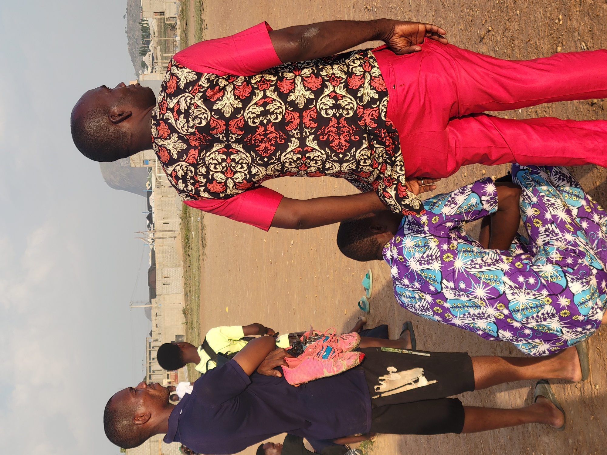 The image depicts several individuals in an open, undeveloped area near what appears to be the outskirts of Ibo, Nigeria. One man, wearing a brightly colored red suit and a floral-patterned shirt, stands with his hand on the shoulder of another individual wearing a patterned purple shirt. Another man is wearing a blue shirt, and the remains of a pair of shoes can be seen. Other individuals are visible in the background. Construction can be seen in the background. The ground is dirt or gravel.