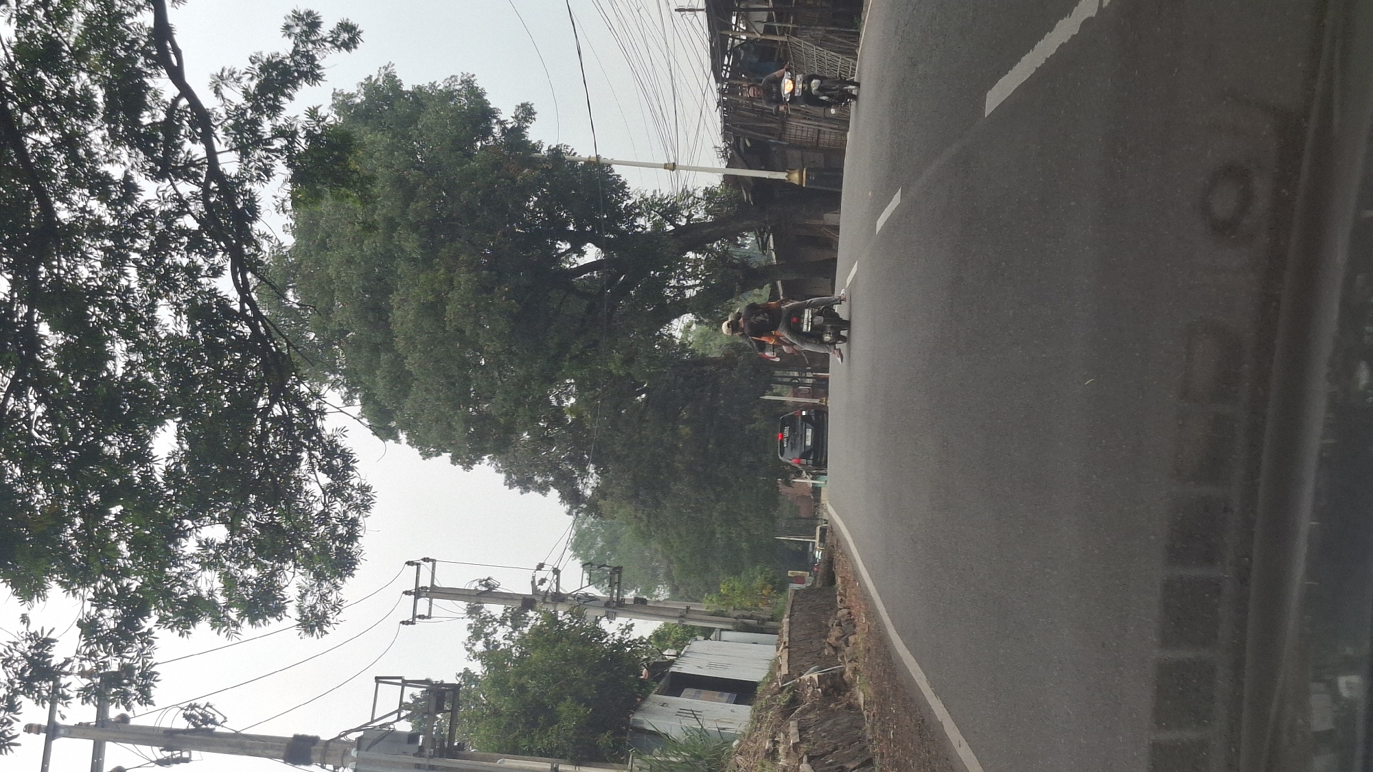 The image depicts a street scene in Cilaku, Indonesia. In the foreground, an asphalt road with white lane markings runs towards the horizon. On the right side of the road, a person is riding a motorcycle. Further down the road, two people are riding on a motorcycle in the same direction. A black car is also visible on the road, driving in the same direction. To the left of the road, there is a sidewalk with a stone retaining wall and some buildings. Utility poles with electrical wires are present along the side of the road, and large trees with dense foliage line the street, some obscuring the sky. The sky is overcast, suggesting a cloudy or hazy day. The overall environment appears to be a suburban or semi-rural area.