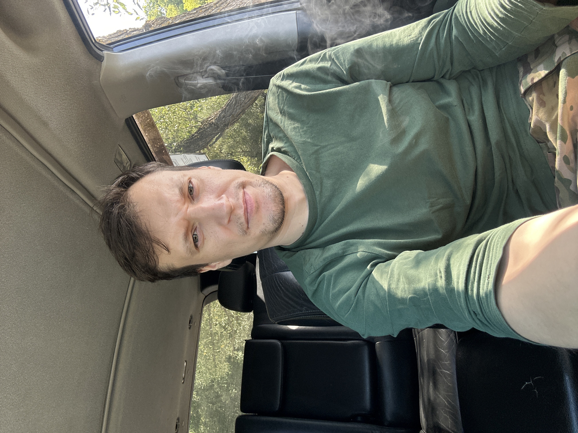 A man is sitting in the driver's seat of a vehicle, likely a car or SUV, with a visible overcast ceiling of the car. He is looking directly at the camera, with a neutral expression. He has brown hair and is wearing a green long-sleeved shirt and camouflage pants. The interior of the car appears to be dark leather with some wear. Through the open window on the right, trees and foliage are visible, suggesting an outdoor location. There is a thin plume of smoke or steam rising from the right side of the frame, near the window. The overall lighting suggests it is daytime. The location is identified as Makiv, Ukraine.