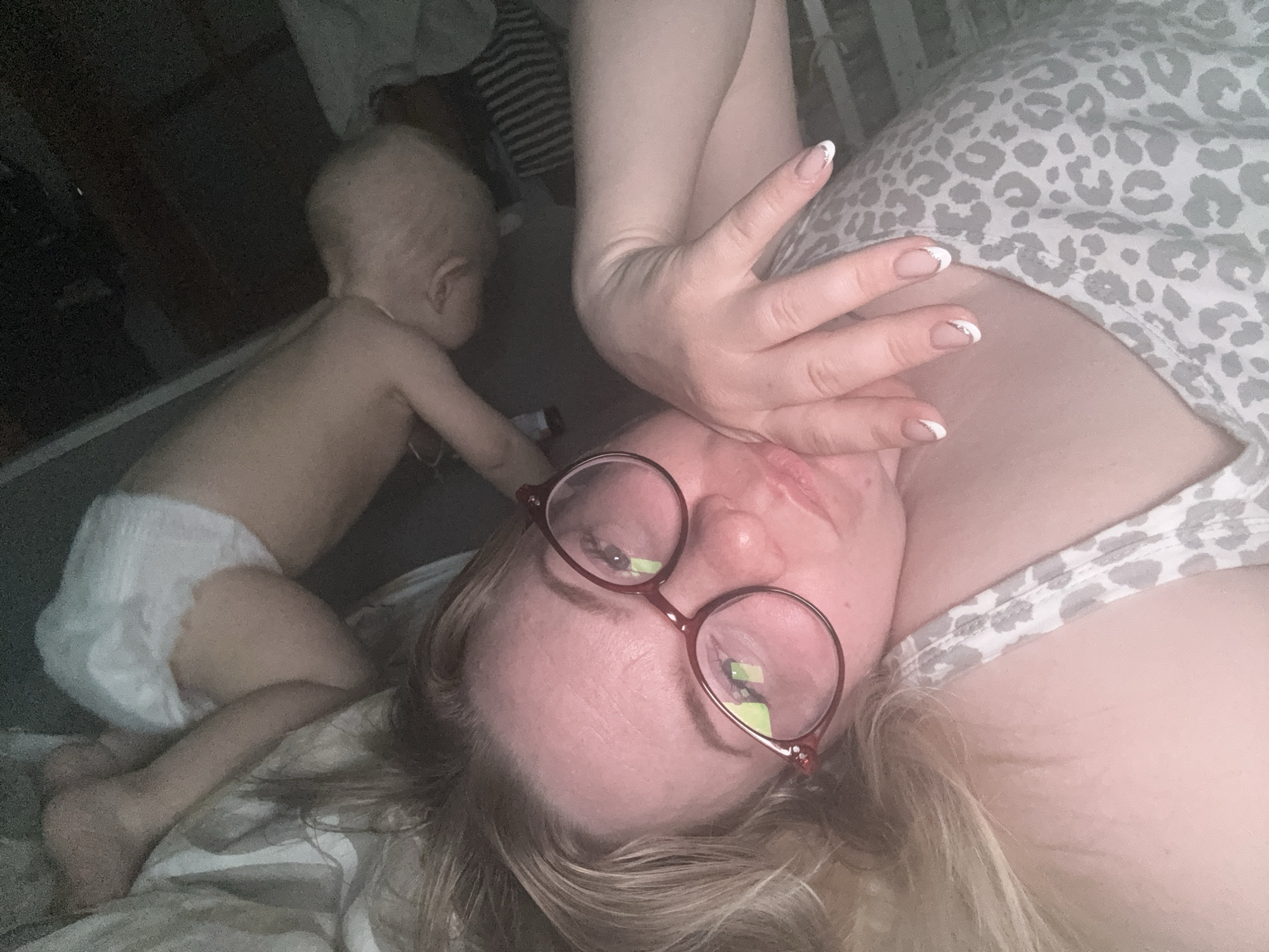 A selfie-style photograph features a woman lying down, facing the camera, with a baby in the background. The woman wears eyeglasses with a red frame and has blonde hair. Her left hand is raised near her face, showing manicured nails with white tips. She wears a gray and white animal-print top. In the background, a baby wearing a white diaper is crawling on the floor. The location is in Chaikovsky, Russia. The environment appears to be a bedroom. The woman's expression is somewhat tired or weary. The baby is focused on something out of view. The scene is domestic and candid.