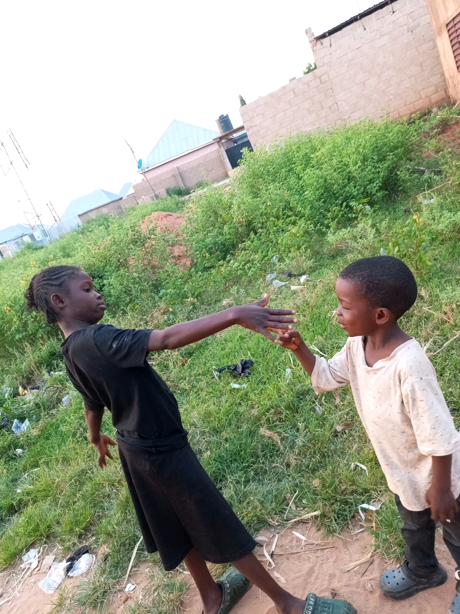 In Tempure, Nigeria, two young children, a girl and a boy, are engaged in an interaction outdoors. The girl, on the left, is wearing a black t-shirt and skirt. She has her right arm extended, with her hand open, palm facing outward, as if offering or accepting something. Her hair is styled in braids. The boy, on the right, is wearing a light-colored, long-sleeved shirt and dark pants. He is looking at the girl's hand and appears to be reaching out to it with his own hand. They are standing on a sandy area with patches of green grass and some scattered debris, including plastic bags. In the background, there are brick buildings with corrugated metal roofs, some construction materials, and dense green vegetation. The sky above is bright and overcast.