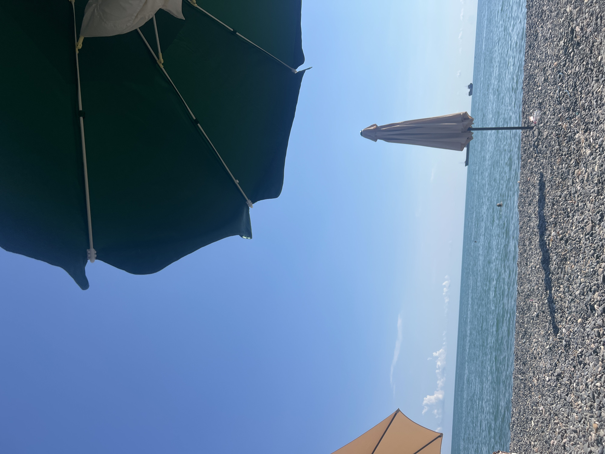 A wide shot shows a pebble beach with the sea and sky in the background. Two beach umbrellas are visible, one a closed, light tan umbrella standing upright in the center, and a large, dark green open umbrella dominating the upper right quadrant of the image. A portion of another open, tan umbrella is visible in the upper left corner. The sea is a calm blue with a faint horizon line where it meets a clear blue sky. A few wispy clouds are present near the horizon. Two ships are visible in the distance on the water. The pebble beach is densely covered with smooth, gray, and dark stones. Shadows are cast across the beach by the umbrellas. The scene suggests a tranquil day at the seaside in Sokhumi, Abkhazia.