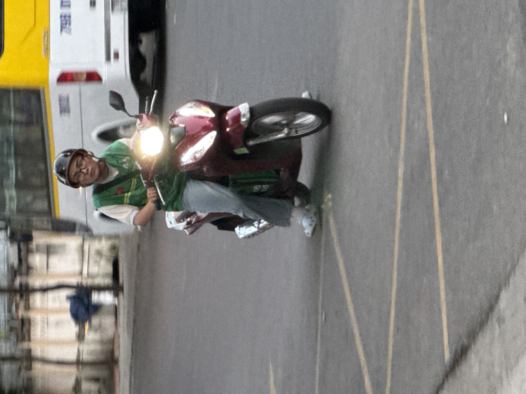 A person wearing glasses and a helmet rides a maroon scooter on an asphalt road. The person is wearing a green vest over a light-colored shirt and grey pants. The scooter's headlights are on. In the background, a yellow and white bus is visible, along with some buildings and trees. The road has yellow lane markings. The scene appears to be an urban environment in Nha Trang, Vietnam.