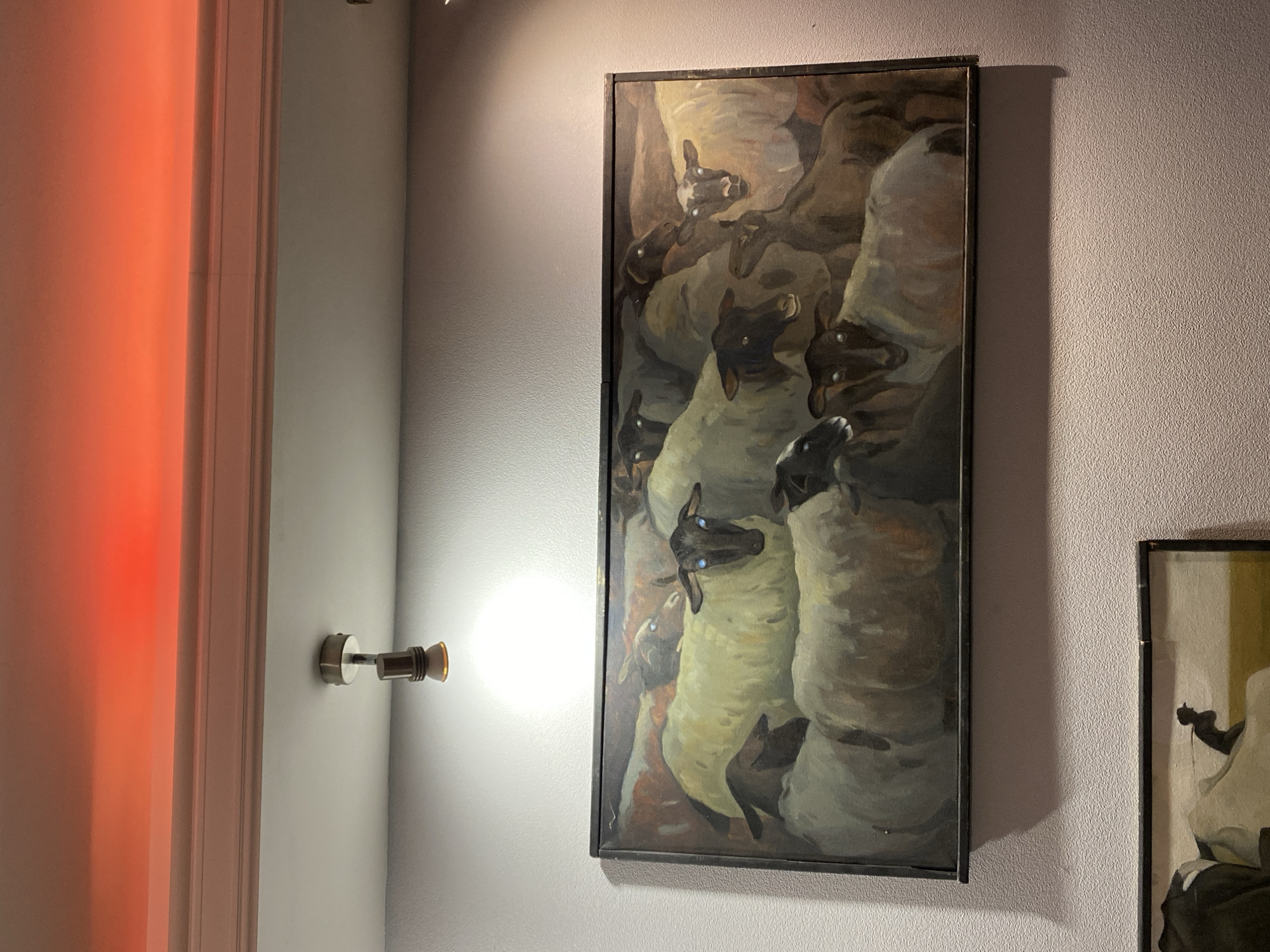 A close-up, indoor shot presents two framed artworks hanging on a light purple textured wall. The left frame is taller and narrower, showcasing a painting dominated by numerous sheep with dark faces and blue eyes, painted in shades of off-white, brown, and gray. The sheep are tightly packed, creating a dense composition. The frame is dark brown or black.
To the left of the taller artwork is a light fixture extending from the wall and a door frame painted in white that is bathed in red light. A portion of another artwork is visible on the right, framed in dark wood with a matte golden image. The wall has a subtle texture. The scene does not appear to be set in Moscow, Russia. There are no people visible in the frame.