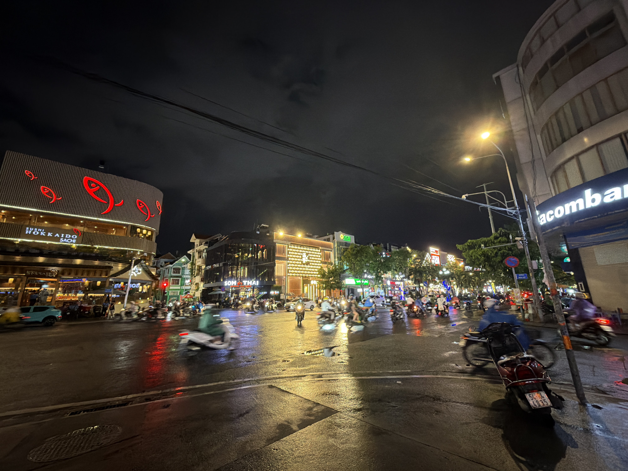 The image depicts a busy intersection at night in Thủ Đức City, Vietnam, likely on a wet street given the reflections. Several motorcycles and a light blue car are visible, some in motion, indicating traffic. Buildings line the street, illuminated by various lights, including neon signs and streetlights. One prominent building features a sign that reads "SUSHI HOKKAIDO SACHI" with red fish motifs. Another building has a large, brightly lit sign with the name "acombank." The sky is dark and cloudy, typical of a night scene. The overall atmosphere suggests a lively urban environment with active transportation and commercial activity.