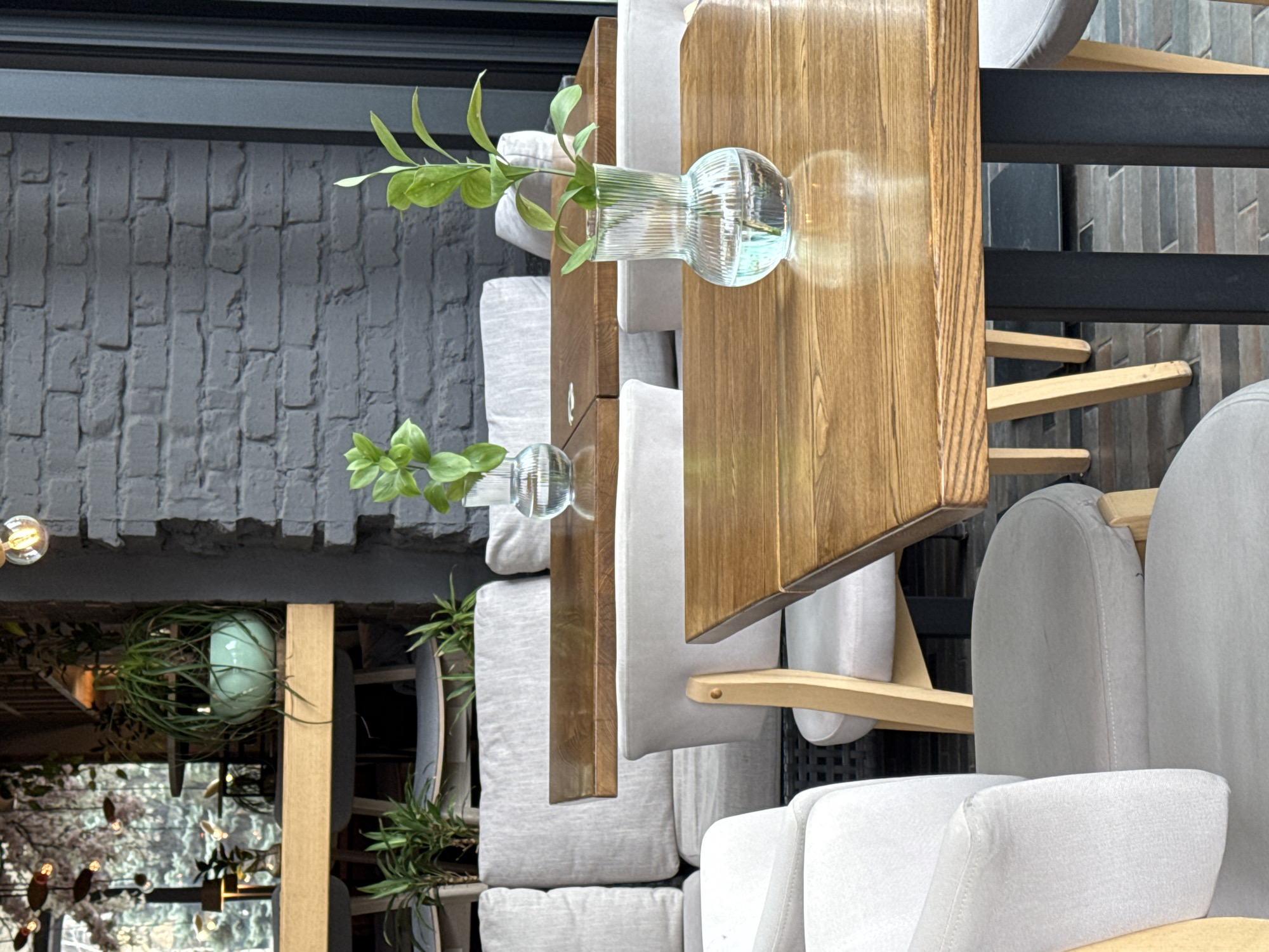 The image captures an indoor scene, likely in a restaurant or café with a stylish, modern design. Visible is a gray brick wall in the background. There's a set of wooden tables with light gray cushioned chairs. On the tables are clear glass vases containing green plants or cuttings. In the lower left corner, a hanging plant and lights can be seen, adding to the ambiance. The scene is well-lit, with a mix of natural and artificial lighting. The overall aesthetic appears to be clean, contemporary, and plant-filled. The exact location within Unknown, Unknown cannot be determined with the given information.