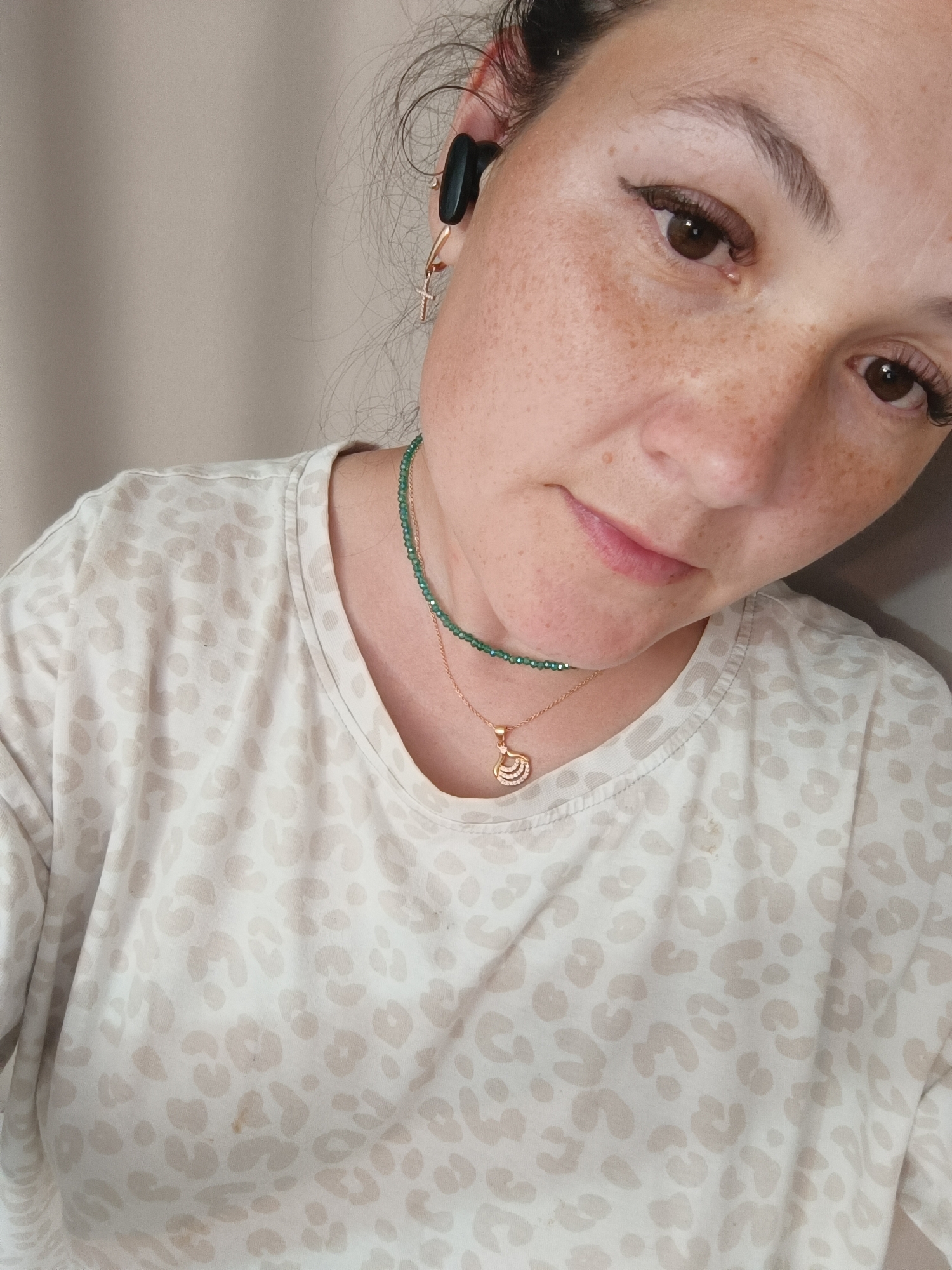 The image is a close-up, eye-level, selfie-style shot of a woman in Chaikovsky, Russia. She has light brown hair, brown eyes, and fair skin with freckles. She is wearing a light-colored t-shirt with a leopard print pattern, a green beaded necklace, and a rose gold necklace with a shell-shaped pendant. She also has on black earbuds and a small gold cross earring. The background is a neutral beige wall. The woman is looking down and slightly to her right, with a neutral expression. The image is well-lit, with natural light coming from the left.
