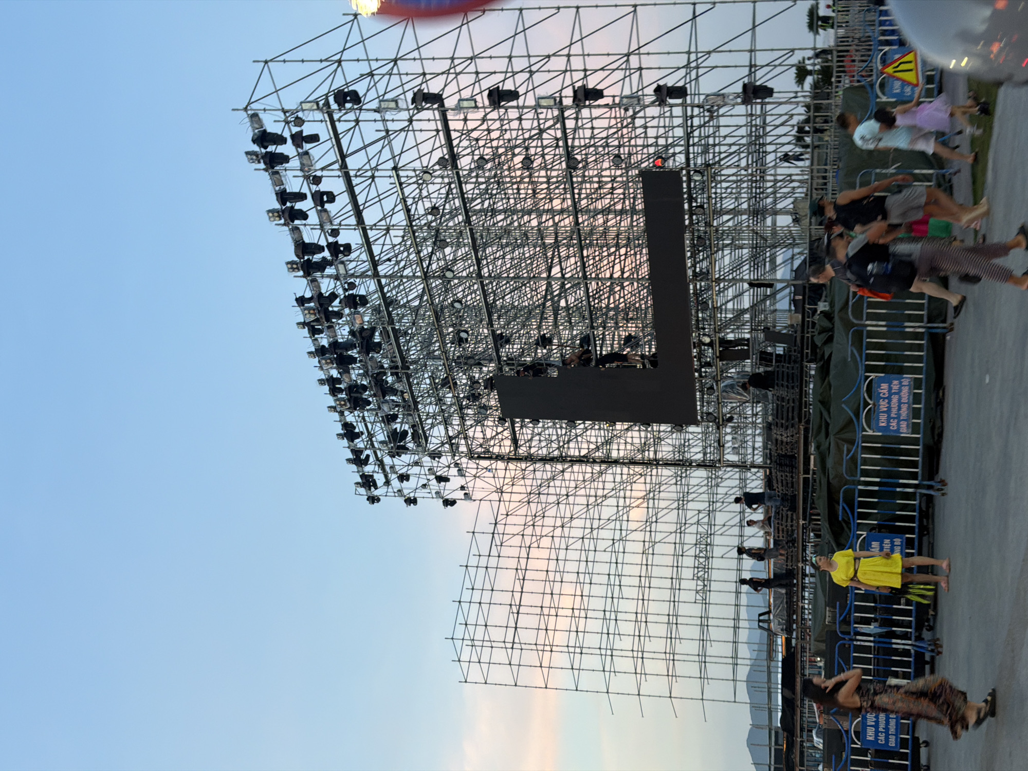 The image depicts an outdoor scene in Nha Trang Ward, Vietnam, likely during dusk. A massive scaffolding structure, resembling a large stage or event setup, dominates the background. This structure is adorned with numerous lights and appears to be constructed from metal poles and frames. A large, dark rectangular screen is visible within the scaffolding, with a distinct "L" shape integrated into its design.
In the foreground, several individuals are present. To the left, a woman in a patterned dress walks away from the camera. Near the center, a woman in a bright yellow dress stands, holding a bag. Further to the right, a group of people, including a child in a purple dress, are walking. The ground is paved, and blue metal barriers are set up, with signs bearing Vietnamese text. In the far background, palm trees and distant hills or mountains are visible, suggesting a coastal or scenic location. The sky is a soft gradient of blue and pink, indicative of twilight.
