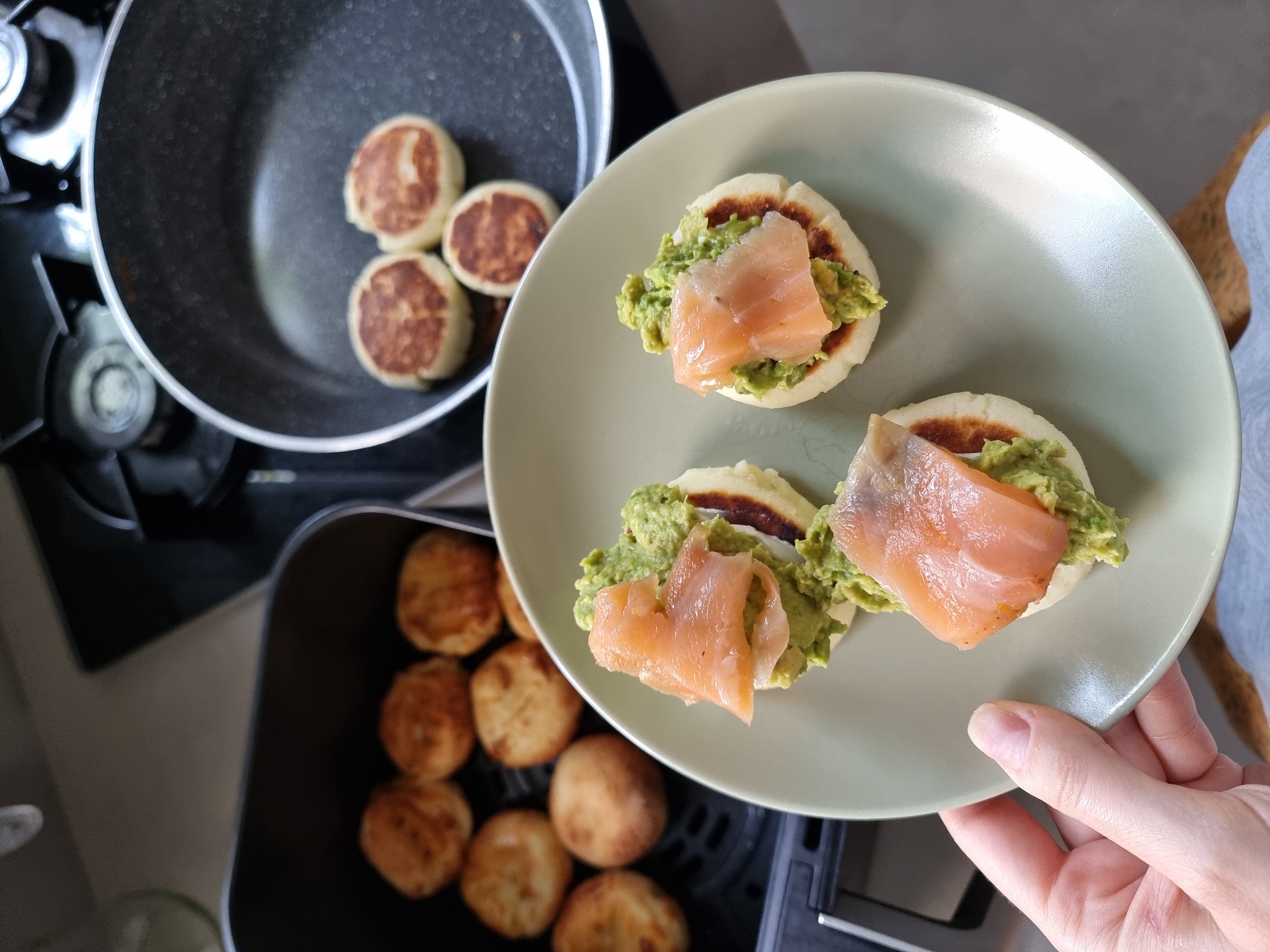 The image displays a top-down view of food preparation in a domestic kitchen, likely located in Ramat Gan, Israel. In the foreground, a person's hand holds a light green, round plate presenting three finished servings of a meal. Each serving features a small, round, lightly browned flatbread or patty, topped with a generous dollop of mashed green avocado and a folded slice of light pink smoked salmon.
To the left of the plate, in the mid-ground, a black frying pan is visible on a stovetop with gas burners. Inside the pan, four more similar light-colored, round patties are cooking or cooling, showing browned undersides.
In the lower right portion of the image, a black air fryer basket is slightly pulled out, revealing several more golden-brown, round patties, suggesting they have been cooked or are being kept warm.
The scene conveys an ongoing food preparation activity, with some items still cooking and others already plated for consumption. The lighting is bright and typical of an indoor kitchen. No text is visible in the image.