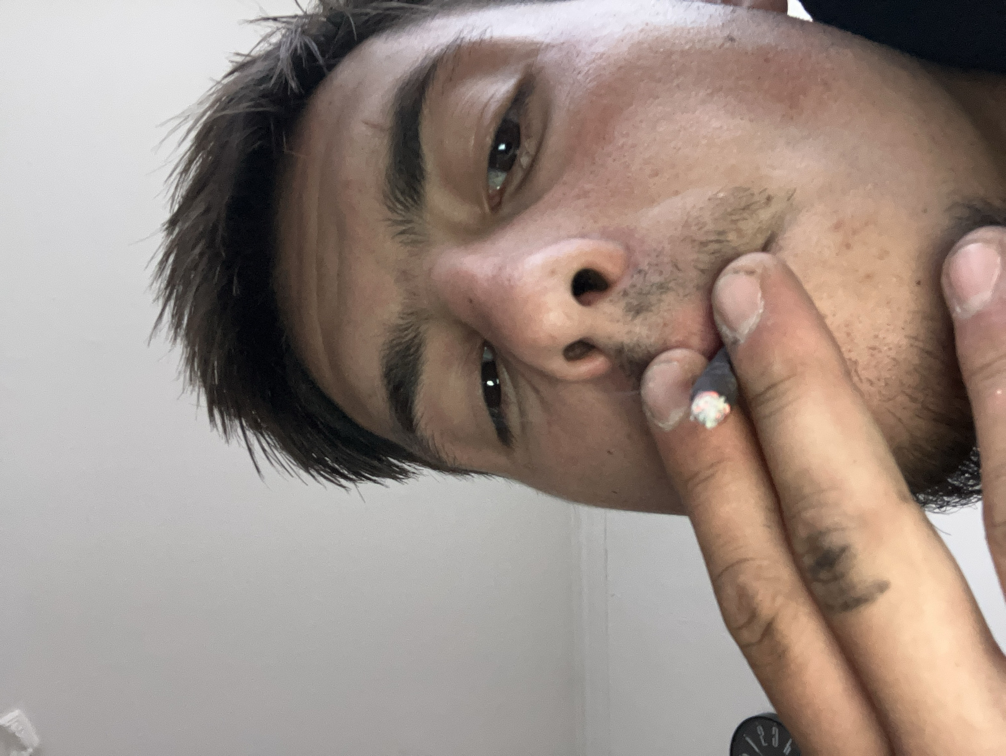 A close-up, diagonally oriented image shows a young person with dark, messy hair and stubble, looking directly at the camera. The subject holds a lit cigarette or joint to their mouth with their right hand.
The visible features include one eye with a dark iris, a distinct eyebrow, and part of the nose and cheek. The skin is medium-toned, with some redness and stubble present around the chin and upper lip area. The fingers holding the lit item appear smudged with a dark, possibly grimy residue, especially on the index finger. The fingernail of the index finger has a unique colorful, glittery embellishment.
The cigarette or joint has a glowing ember and white ash at its tip, suggesting it is actively being smoked. The wrapper appears dark. The background is a plain white wall, indicating an indoor setting. A small, dark object is partially visible in the bottom right corner, showing faint, largely illegible markings that resemble numbers, possibly "1/23". The lighting is soft, likely indoor, and the overall mood is candid. The location context is Plovdiv, Bulgaria, though no specific visual cues within the image confirm this.