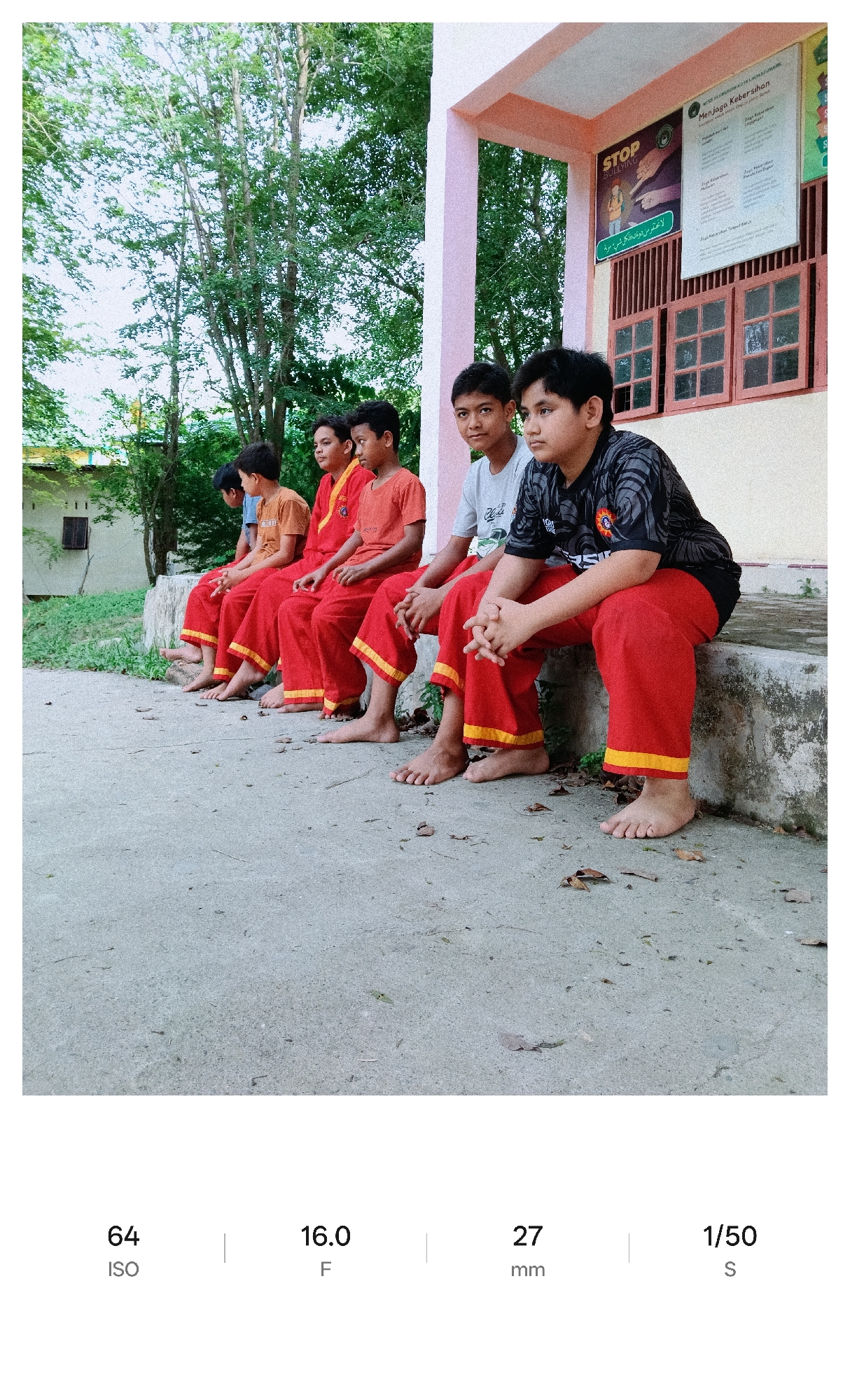 The image shows a group of five young males, likely students, sitting side-by-side on a concrete ledge or bench outside a building. The students are dressed in matching red pants with yellow stripes near the bottom hems, with the exception of the student on the right who is wearing a black patterned shirt and the second one from the left who is wearing a red outfit. Each student is barefoot. The building has pink trim, and visible on the wall behind the students is a sign with the word "STOP BULLYING" and another sign titled "Menjaga Kebersihan" which translates to "Maintain Cleanliness." The background includes trees. The image was taken in the city of Lhokseumawe, Indonesia. Camera settings indicated at the bottom of the image state ISO 64, F/16.0, 27mm, and 1/50 second.