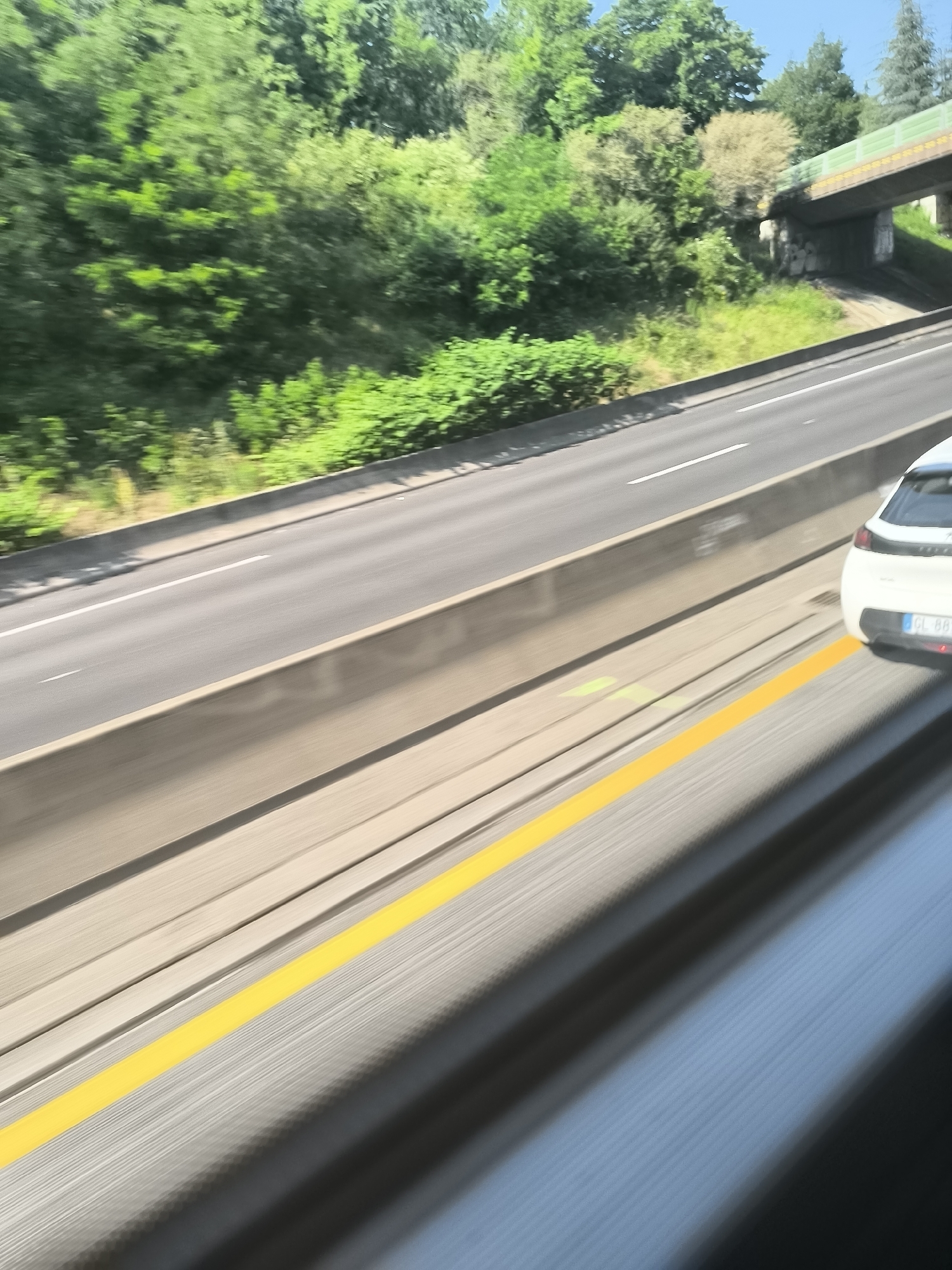 A blurred, angled view from inside a moving vehicle shows a multi-lane highway near L'Horme, France. A white car is visible in the right lane. A concrete barrier separates the lanes from a vegetated area. An overpass is also visible. A yellow line is painted on the side of the highway closest to the camera.