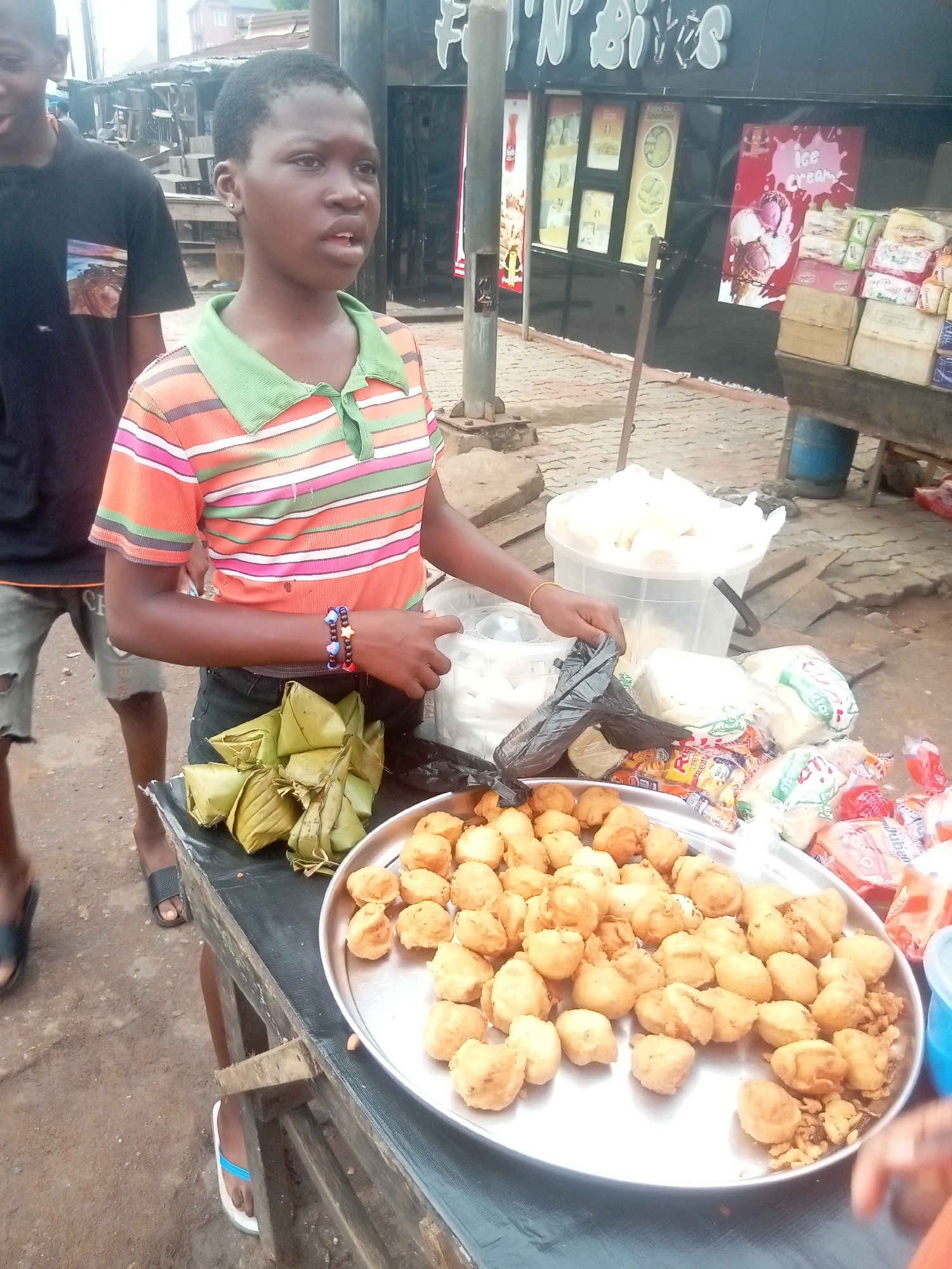 A young person stands behind a table displaying food items in Nkpor, Nigeria. The individual is wearing a striped orange, white, and green shirt. They have two bracelets on their left wrist. The table holds a large, round, silver platter filled with fried dough balls. Next to the platter are triangular packages wrapped in green leaves, a clear bucket of white contents, and various wrapped items.
A person wearing a black t-shirt and shorts is standing to the left of the table. The background features a building with the sign "FUN 'N' BITES" visible, and an ice cream advertisement is displayed. The surrounding environment appears to be an outdoor market or street vending area.