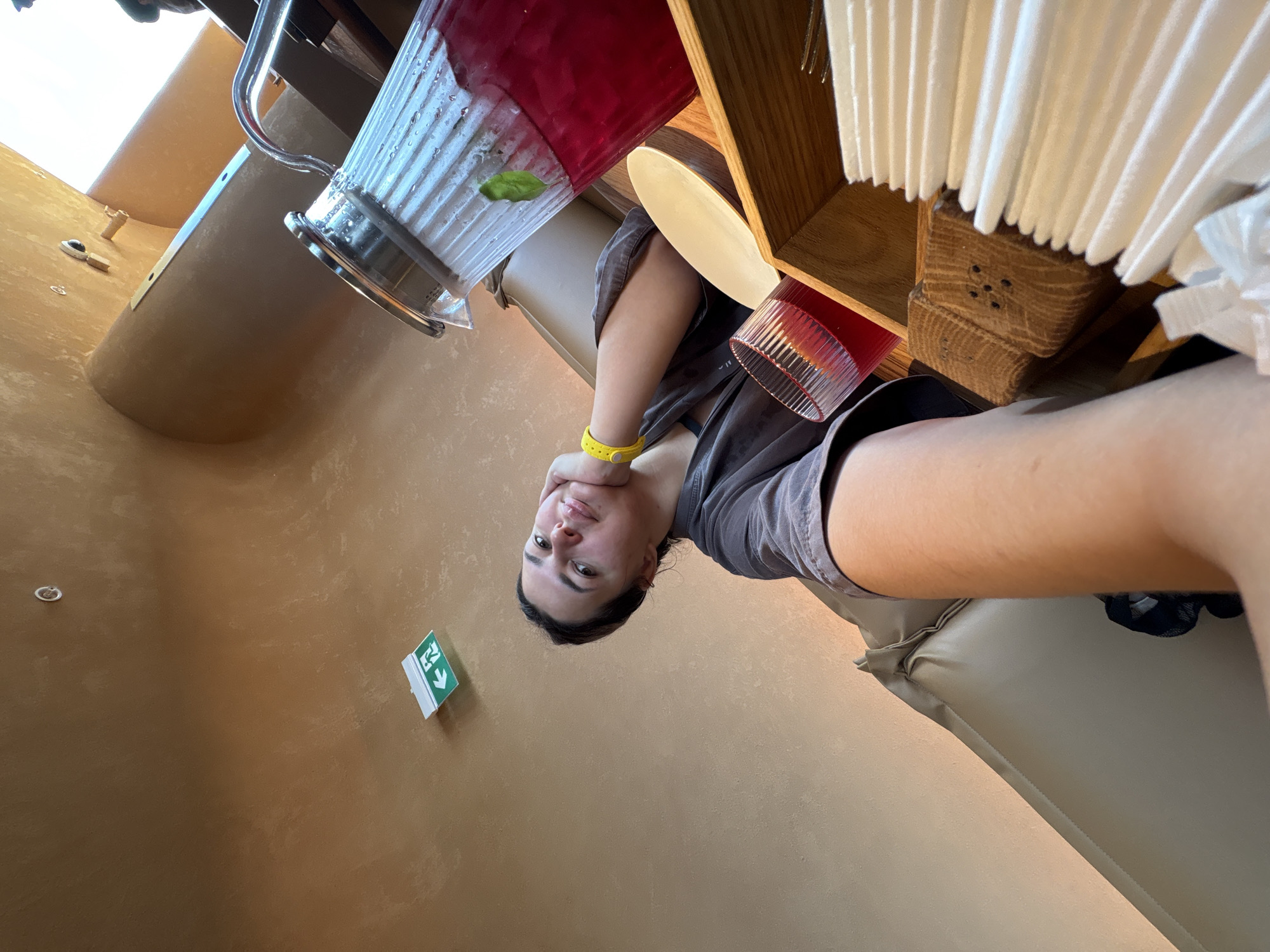 A young woman with dark hair is taking a selfie in a dimly lit, modern room with a muted brown, textured ceiling and walls. She is wearing a dark grey, loose-fitting top and a bright yellow wristband. Her left hand is resting on her chin as she smiles softly at the camera. In the background, a large glass pitcher filled with a red liquid and a sprig of mint, a small glass with red liquid, a white plate, and a wooden holder filled with white napkins are visible on a wooden surface. To the left, a portion of a beige leather sofa is visible. An illuminated green exit sign with an arrow pointing left is mounted on the wall above and behind the woman. The overall ambiance suggests a spa or a lounge area. The location is not specified in the image itself and cannot be determined from the visual information.