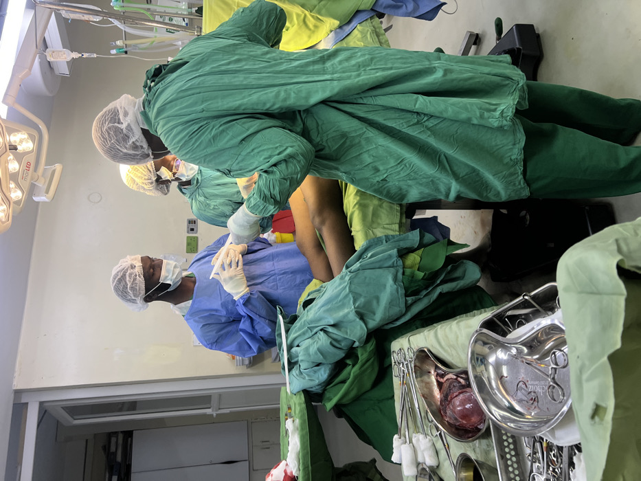 An operating room scene in Nairobi, Kenya, shows three medical professionals engaged in a surgical procedure. The room is brightly lit by overhead Maquet LED surgical lights. A patient lies supine on a draped operating table, with their lower extremities, specifically the upper thigh/groin area of the right leg, exposed.
Of the three individuals, one person in blue surgical scrubs, a hair net, face mask, and gloves stands on the left, holding a syringe with both hands. A second individual, wearing green surgical scrubs, a hair net, face mask, and gloves, is positioned with their back mostly to the camera, facing the patient's exposed leg, seemingly performing a surgical action. A third individual, also in green scrubs, a hair net, face mask, and gloves, is partially visible behind the first person, to the right of the patient's head.
An intravenous (IV) drip bag hangs above the patient. On a table in the foreground to the left, multiple stainless steel surgical instruments, including forceps and clamps, are organized in trays. One kidney dish contains a dark red mass, appearing to be excised tissue or an organ. Another kidney dish holds additional small instruments. Green and blue surgical drapes cover the patient and surrounding instrument tables. The walls are light-colored, with visible wall-mounted electrical outlets and what appear to be medical gas ports in the background.