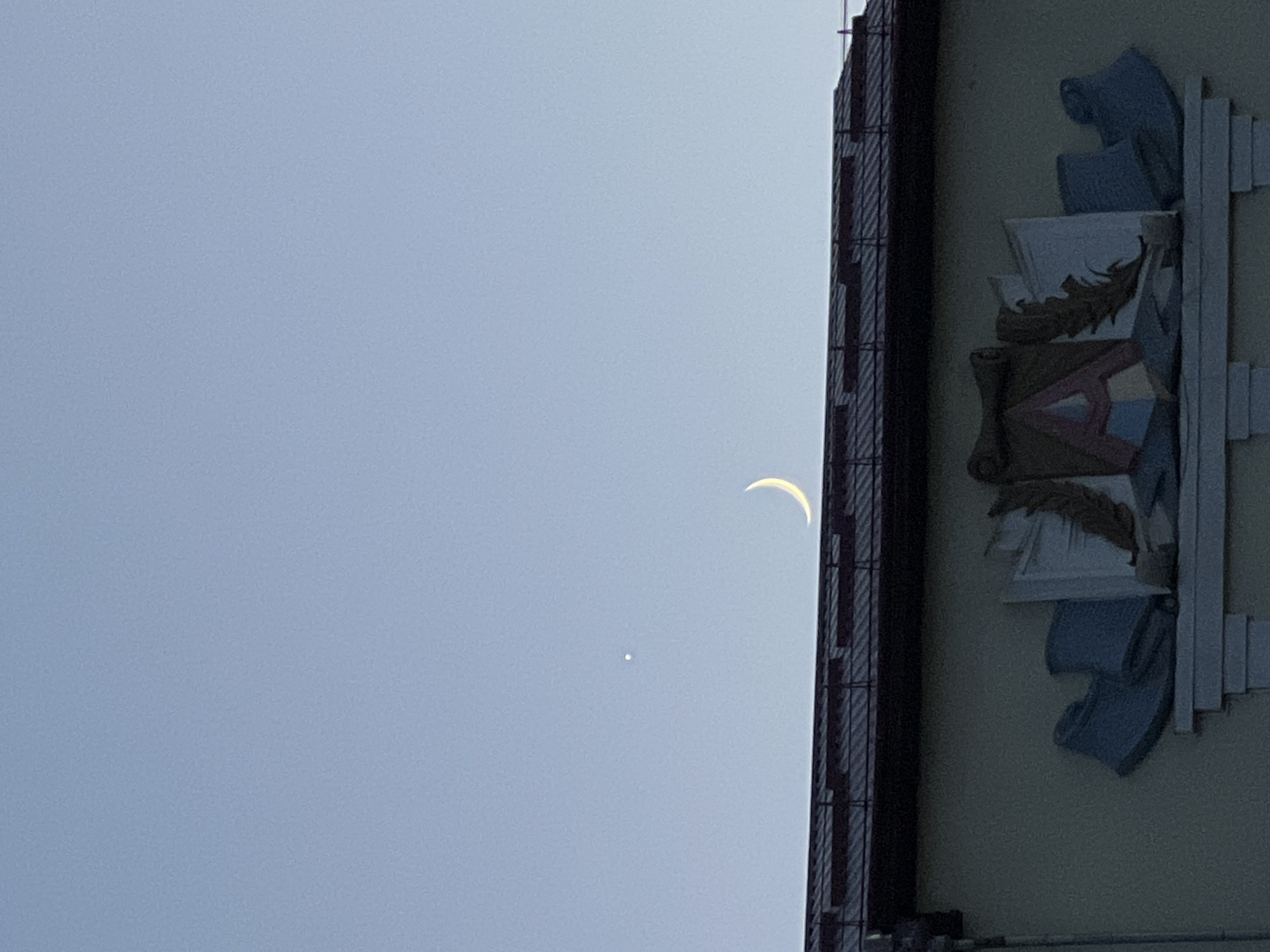 The image captures a clear sky above a building in Ternopil, Ukraine. A sliver of a crescent moon is visible in the upper right quadrant of the sky, while a bright point, likely a planet, is to the left of the moon. The building's roof, with visible shingles, runs across the middle of the frame. Below the roofline, the facade of the building features an architectural relief. This relief includes what appears to be a stylized representation of books, with one large element in the center featuring the letter "A" and a feather motif. To the left and right of this central element are abstract shapes resembling scrolls or folded fabric. The color palette of the relief is muted, with tones of beige, blue, and red. The overall lighting suggests either dawn or dusk.
