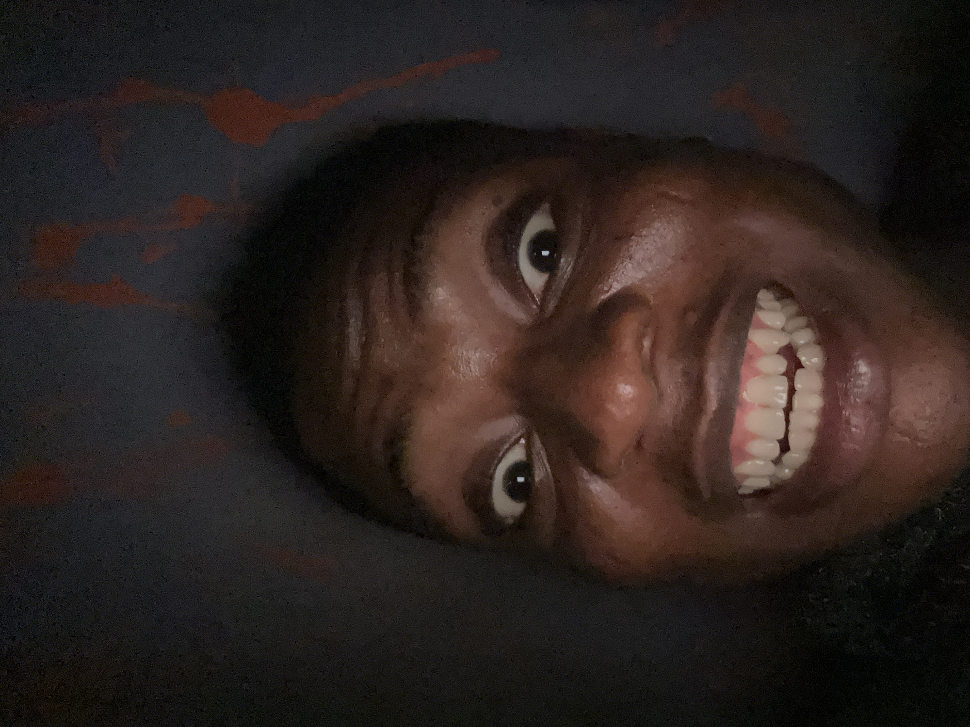 A close-up, low-angle shot shows a person's face with a wide smile revealing their teeth. The person has dark skin, dark eyes, and short dark hair. The background appears to be a dark fabric with some faint red patterns. The location is not specified and the details are limited.