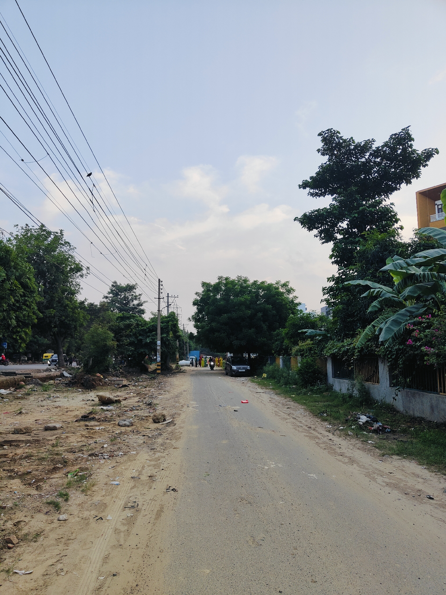The image depicts a street scene in Gurgaon, India. The road is unpaved in some sections and appears to be a two-lane road. To the left of the road, there is a dirt embankment with debris and construction materials. Several trees line the left side of the road, with power lines overhead. A yellow auto-rickshaw and a car are visible on the road in the distance.
On the right side of the road, there is a wall with a metal fence. Behind the wall, there are lush green trees and foliage, including what appears to be banana plants. A building with orange siding is partially visible in the background on the right. A few people are walking on the right side of the road in the distance, and a motorcycle is being ridden towards the viewer on the right side. The sky is partly cloudy. The overall impression is of a developing area with both natural and built environments.