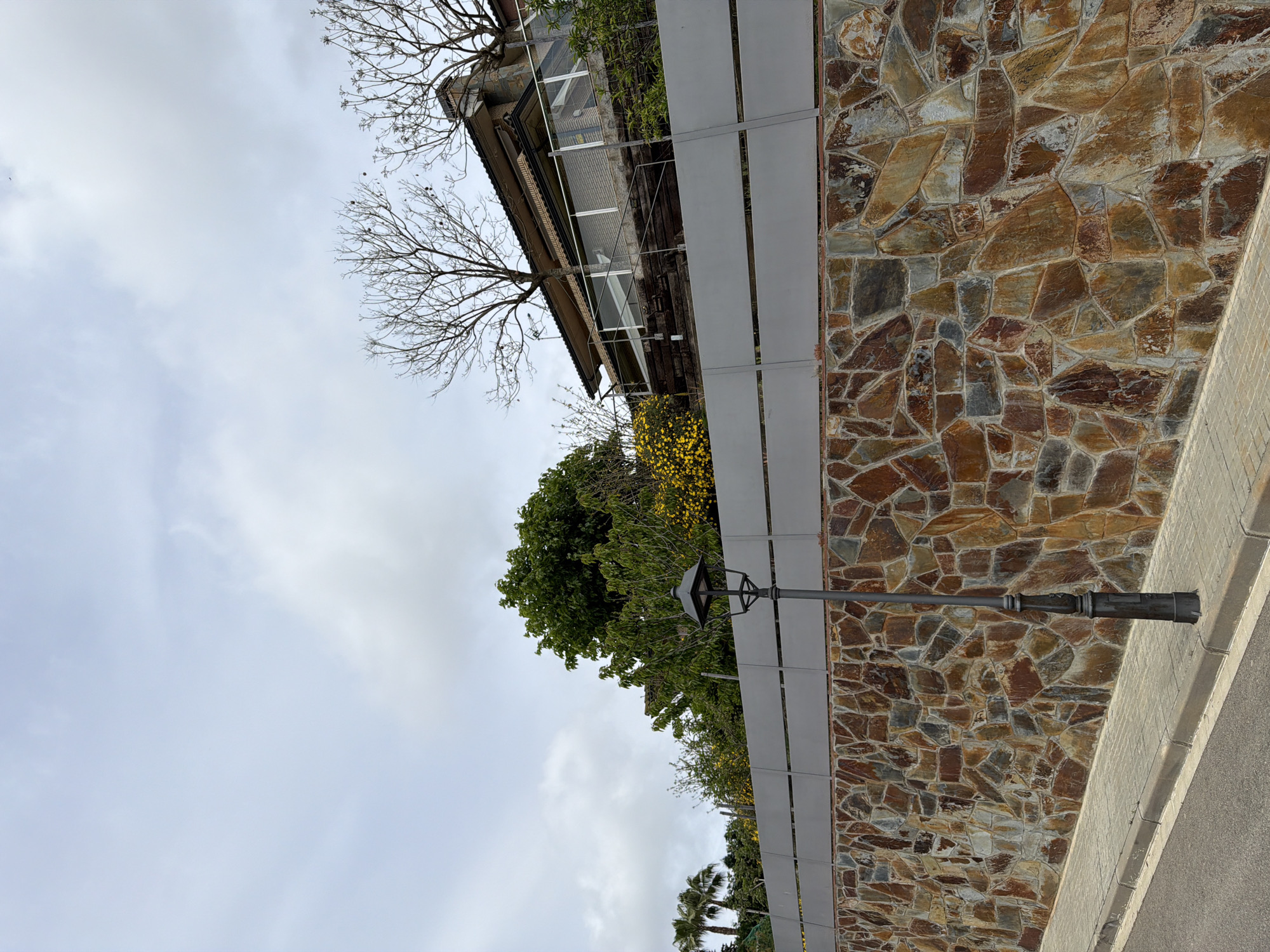 The image depicts a street scene in Sant Andreu de Llavaneres, Spain, featuring a stone wall bordering a road. The wall is constructed of irregular, multi-colored stones and is partially obscured by a grey, metal fence. A streetlight extends from the fence. Beyond the fence and wall are various trees and vegetation. A building is visible in the upper portion of the frame, appearing to be situated on a higher elevation. The sky is overcast with clouds. The image is rotated, with the sky appearing at the bottom and the ground at the top.