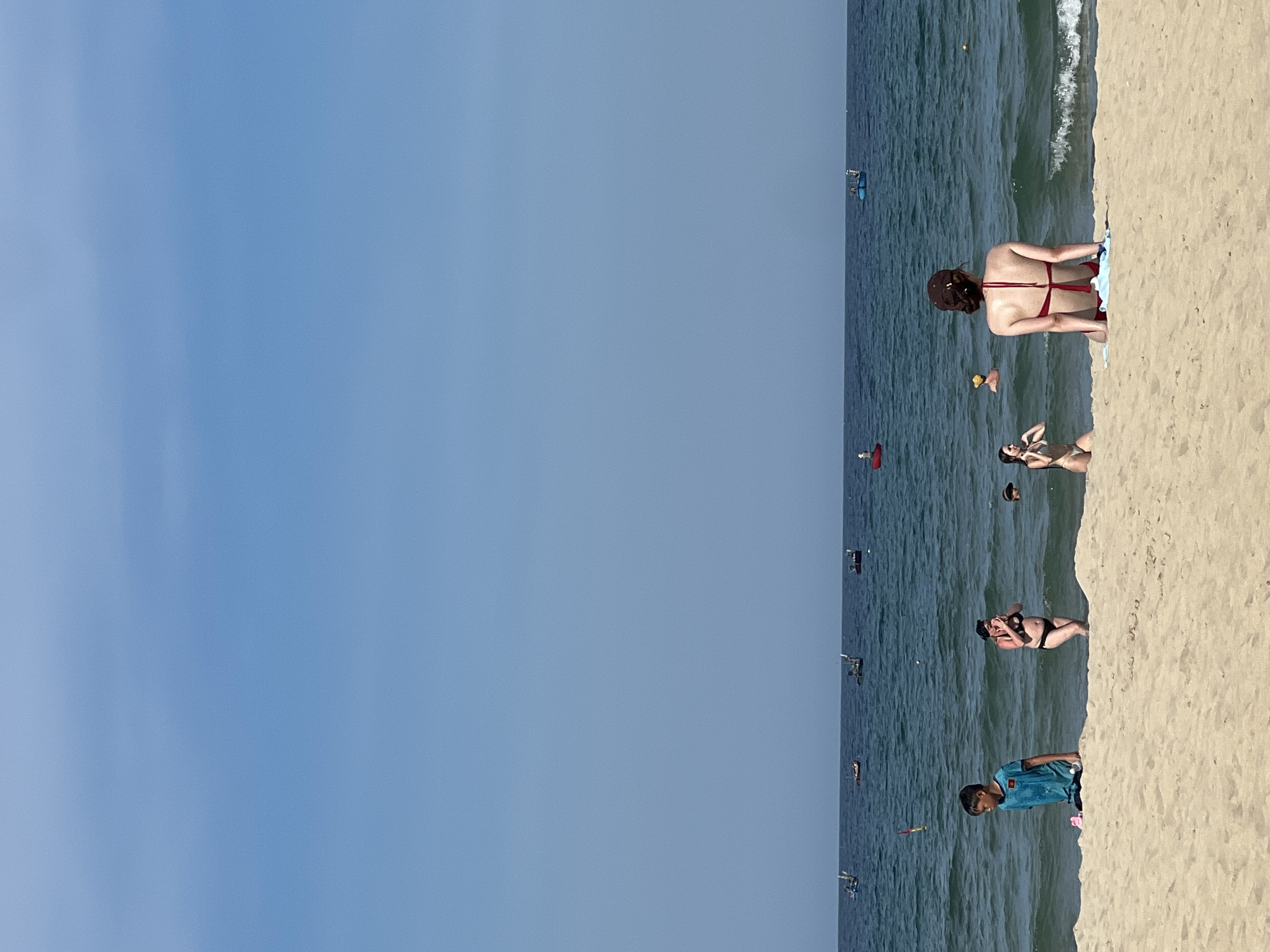 The image depicts a sunny day at a beach in Da Nang, Vietnam. The scene is divided into two main areas: a vast expanse of clear blue sky on the left and the sandy beach and ocean on the right. Several people are visible enjoying the beach and the water.
In the foreground on the sand, a young boy wearing a blue t-shirt is standing. Further back, a woman in a black bikini is lying on the sand. Near the water's edge, a woman in a red bikini is sitting and facing away from the camera. In the ocean, there are several people swimming and wading, with some visible buoys and what appear to be small boats or jet skis in the distance. The water is a deep blue, and small waves are breaking on the shore. The sand is light brown and appears dry.
The time of day is likely afternoon, given the bright sunlight and clear sky. The weather appears to be warm and pleasant, typical for a beach setting in Vietnam. The overall mood is relaxed and recreational, with people enjoying a day at the beach. No visible text is present in the image.