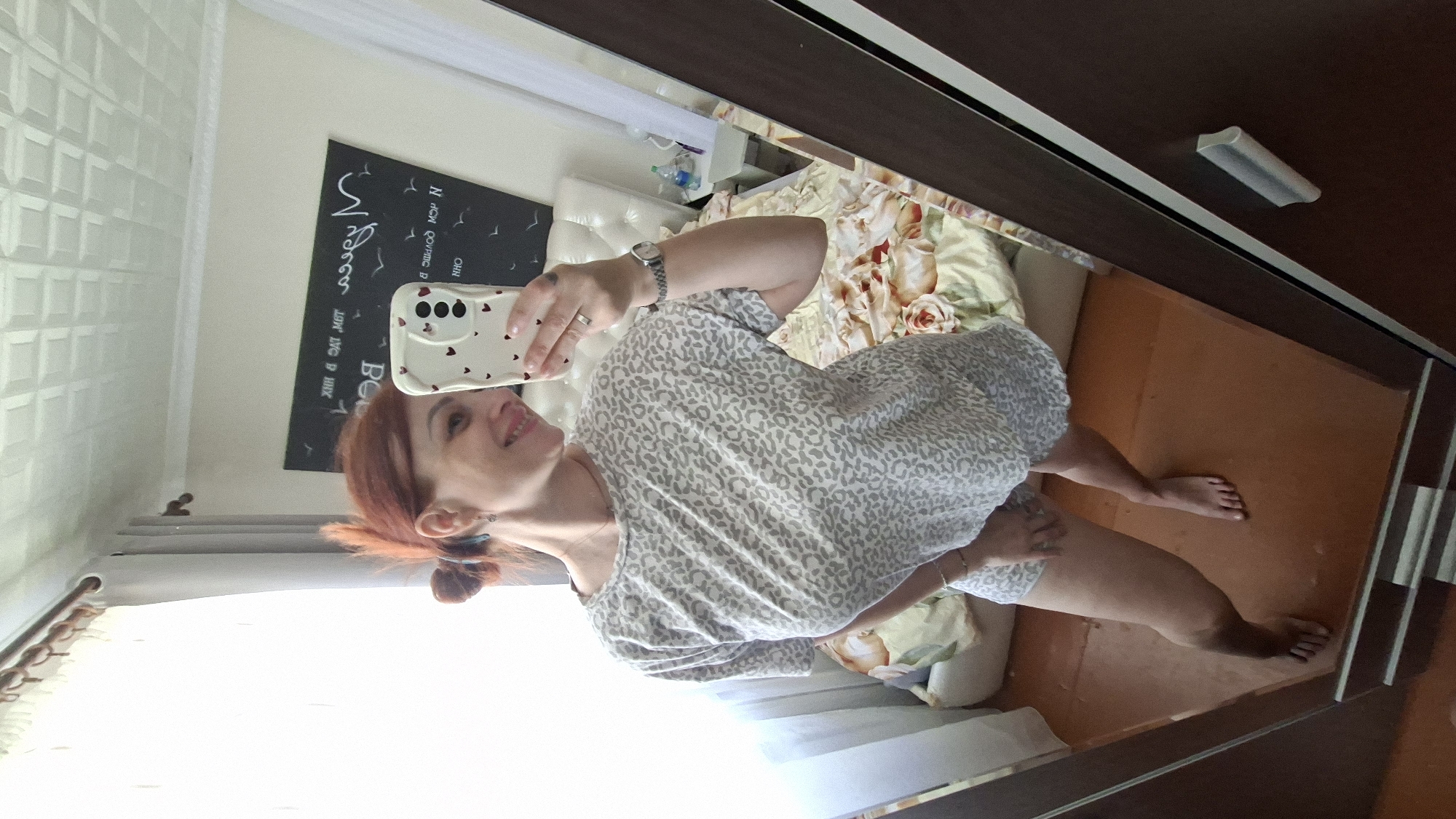 A woman with red hair tied in a bun is taking a mirror selfie in a bedroom. She is wearing a light-colored t-shirt with a gray leopard print and matching shorts. The room features a bed with floral patterned bedding and a headboard. In the background, there is a chalkboard with writing on it, a window with white curtains, and a wardrobe. The woman is holding a smartphone with a heart-patterned case. The floor is a wooden surface.