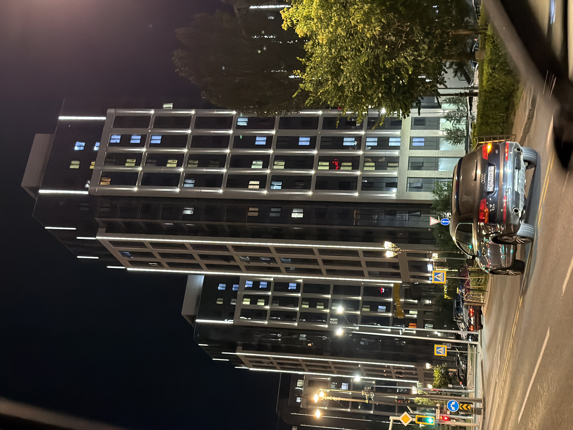 In the city of Astana, Kazakhstan, at night, a dark-colored Audi Q5 SUV drives on a multi-lane asphalt road. The SUV has its license plate visible, reading "609 ARP 01". In the background, two modern, tall residential buildings with prominent vertical white lighting strips are illuminated. The buildings have many windows, some of which are lit with warm yellow and cool blue light. Trees with green foliage are present on the right side of the frame, partially obscuring the buildings. Traffic lights and street signs, including pedestrian crossing signs and a traffic light showing green, are visible along the roadside. Other vehicles, including a red sedan, are parked or driving in the distance. The scene is captured from a low angle, possibly from another vehicle.