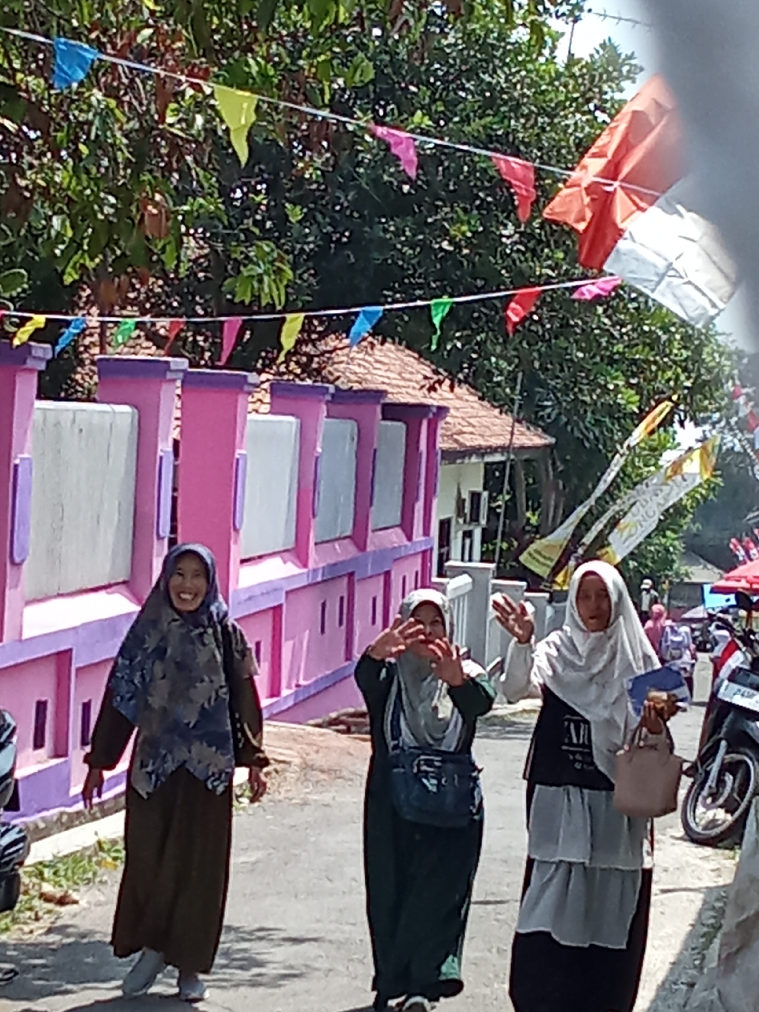 Three women wearing hijabs are walking on a paved street in front of a brightly painted pink fence. The woman on the left is wearing a dark green dress and a blue and grey patterned hijab. She is smiling and looking at the camera. The woman in the middle is wearing a dark green dress and a grey hijab, and she is holding her hands up to her face. The woman on the right is wearing a white hijab and a black and white outfit. She is holding a light brown purse and a blue object. The street is decorated with colorful triangular flags and banners. In the background, there are trees and buildings. There are also motorcycles parked on the side of the street. The scene appears to be a festive occasion. The presence of Indonesian flags suggests a national celebration.