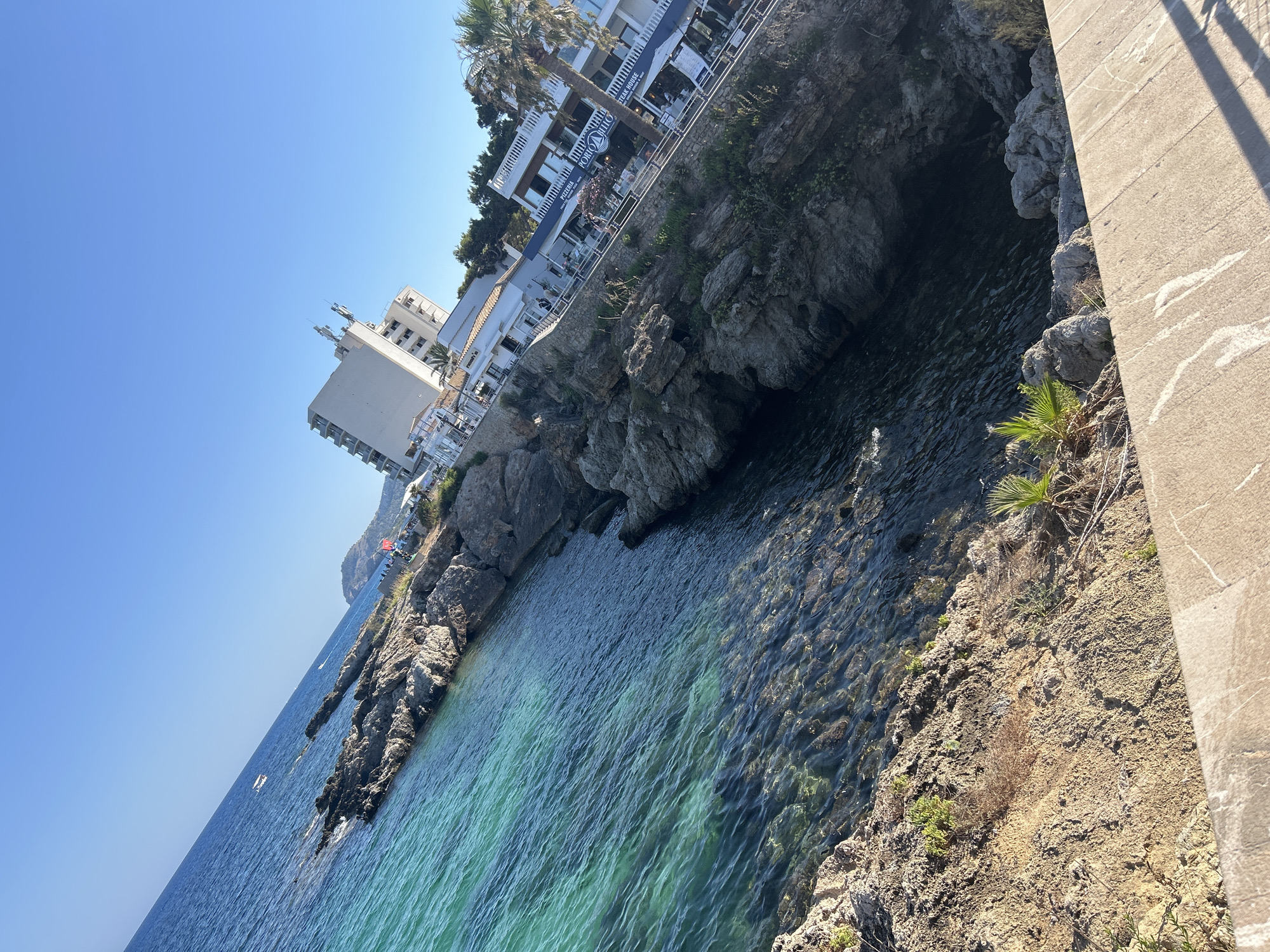 The image depicts a coastal scene in Cala Rajada, Spain, with the sea and rocky shoreline dominating the foreground. The water is clear and turquoise, revealing submerged rocks. On the right side of the frame, a multi-story white building with balconies is visible, situated on the rocky cliffside. Below this building, there are several restaurants with outdoor seating, indicated by tables and umbrellas. One restaurant has signage that reads "PORTO RIO" and "STEAK HOUSE." Palm trees are scattered along the coastline, and the sky is a clear blue. The sun is casting shadows, suggesting it is daytime. A small white boat is visible in the distance on the sea. The perspective of the shot is tilted, giving a dynamic feel to the landscape.