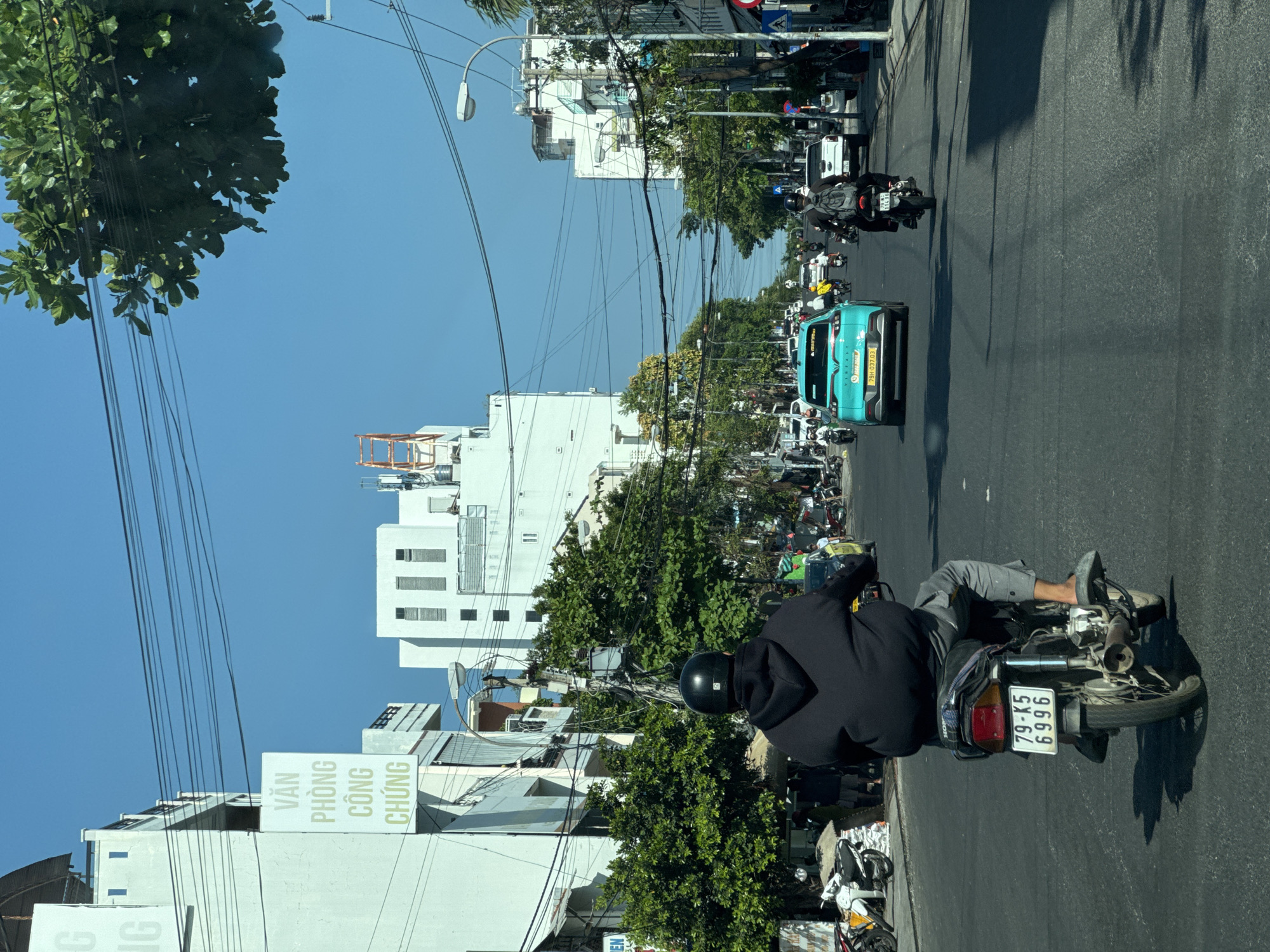 The image shows a street scene in an urban environment, possibly in Vietnam, based on the presence of Vietnamese script. The photo is taken from behind a motorcyclist wearing a black hoodie and helmet. Their license plate reads "79-K5 6996."
In the distance, there's a line of traffic, including a turquoise car with a license plate beginning "79H-037.03" and other motorcycles. Along the sides of the street, there are buildings of varying heights, some of which have signs written in Vietnamese. Overhead are numerous power lines. The sky is a clear, bright blue. The road is asphalt.