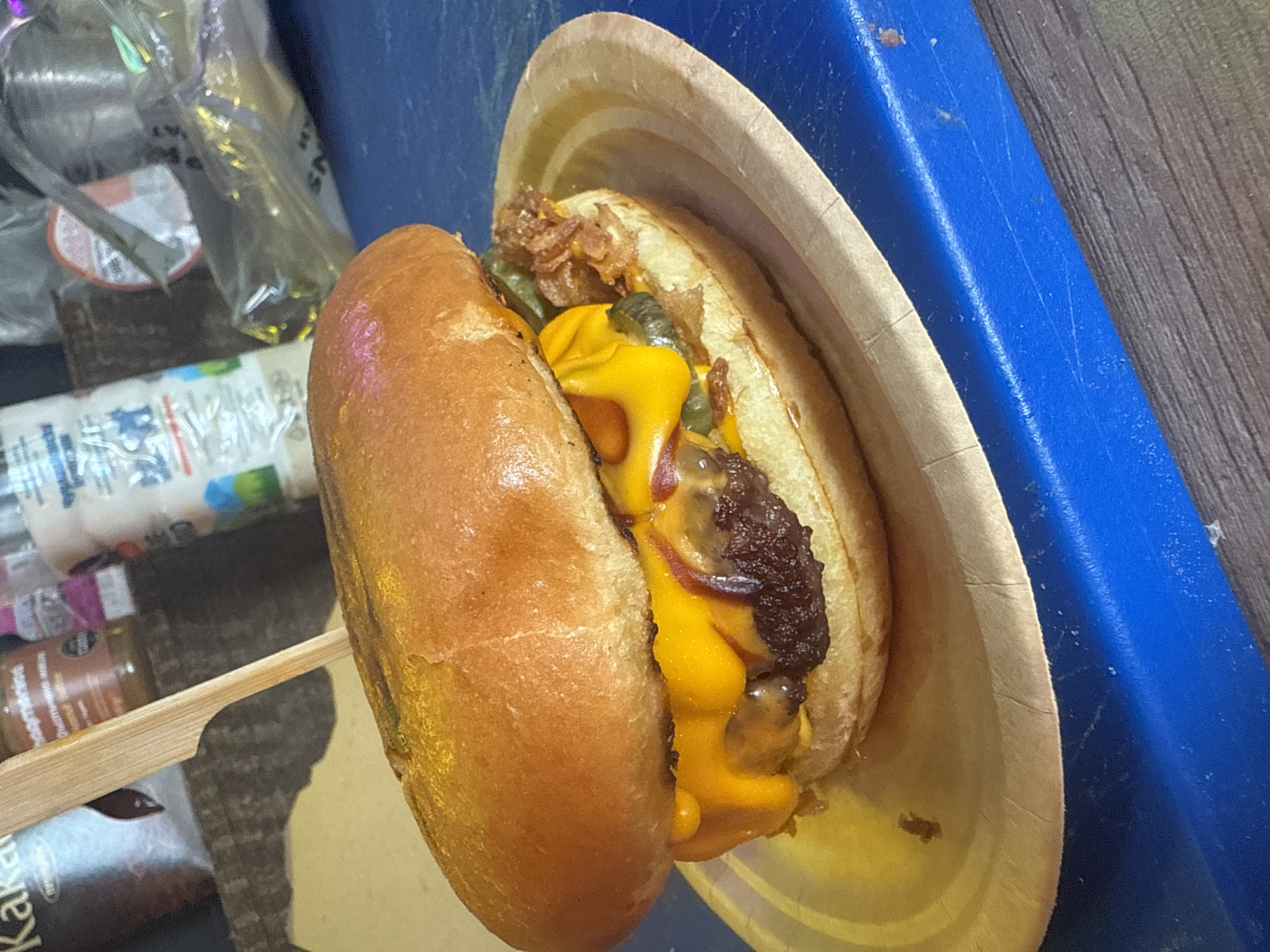 A close-up, diagonal shot captures a prepared cheeseburger-like sandwich resting in a light brown, possibly biodegradable, paper bowl or tray. The sandwich features a beef patty topped with generous amounts of melted orange cheese, green pickled components (likely gherkins or jalapeños), and crispy fried onions, all nestled within a light brown, glossy bun. A wooden skewer is visible, piercing the sandwich to hold it together, with its end extending towards the lower left.
The background is partially blurred but reveals several items. To the left, there are various plastic-wrapped containers and a white bottle with indistinct blue and green labels. Below these items, a brown jar with a label is partially visible, showing "Kaka..." at the bottom left. These items rest on what appears to be a wooden surface. To the right of the paper bowl, a vibrant blue surface, possibly a plastic tray or table edge, is visible, contrasting with the warm tones of the food. The overall lighting suggests an indoor setting. No people are visible.
