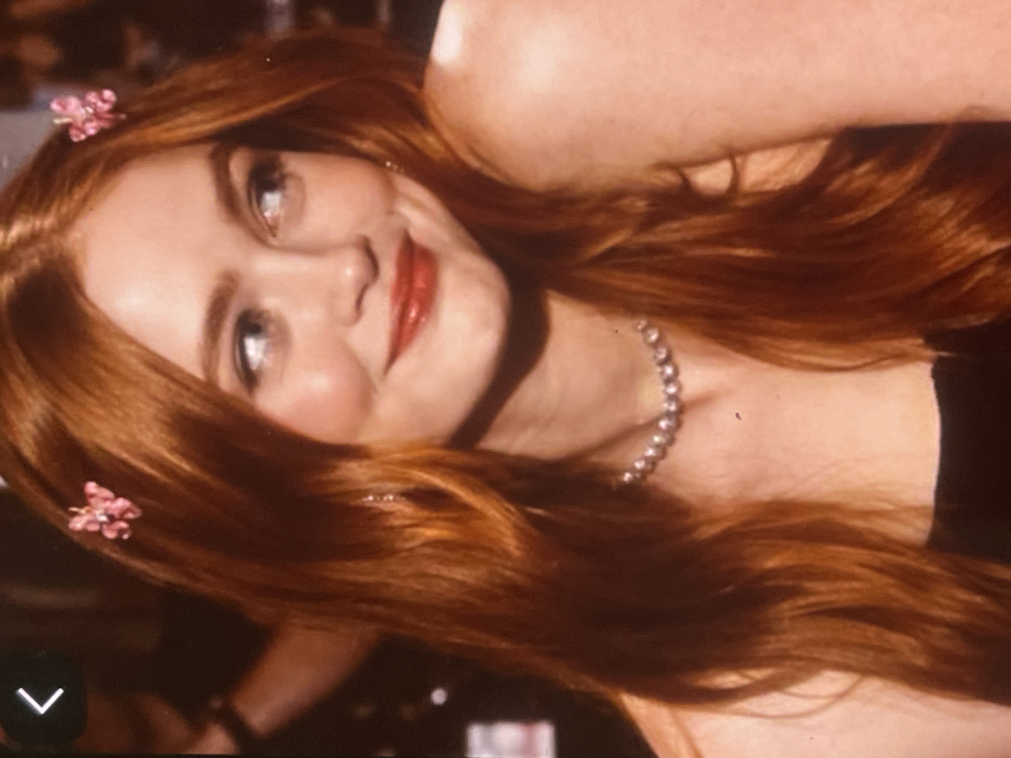 An indoor, tilted portrait features a young woman with long, wavy auburn hair, smiling and looking upwards and slightly to the left. She has fair skin with a blush on her cheeks and is wearing bright red lipstick. Two delicate pink floral hair clips are nestled in her hair, one above her forehead on the left and another on the right side of her head. A string of pearls adorns her neck. Her eyes are a light color. The background is dark and out of focus, suggesting an evening or night setting with soft, warm lighting. A white chevron navigation icon is present in the bottom-left corner of the image. The scene is set in Poltava, Ukraine.