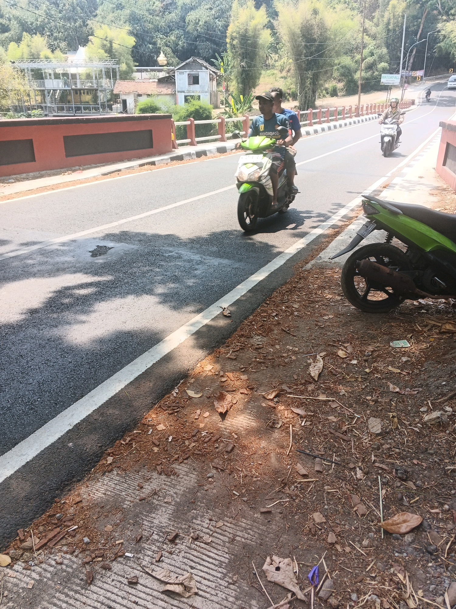 The image shows a road with two motorcycles and their riders. The road is paved and has a white line marking the lane. On the left side of the road, there is a red concrete barrier. On the right side, there is a dirt and debris-covered shoulder with a parked green motorcycle. In the background, there are trees, buildings, and a sign that reads "WATERFALL". The lighting suggests it is daytime. The location appears to be a scenic area, possibly a tourist destination. Based on the provided context, the scene is likely located in Semarang, Indonesia.