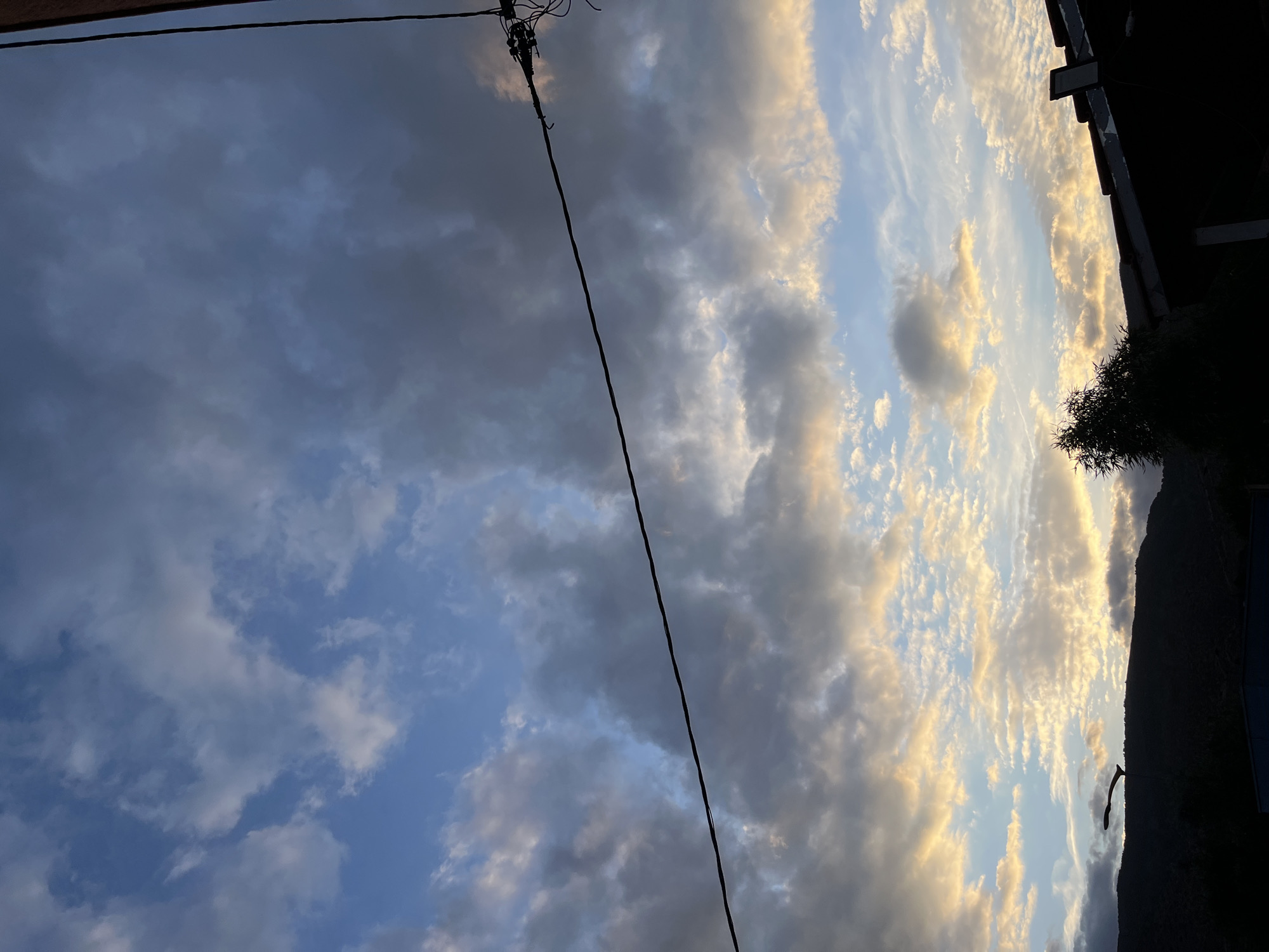 The image shows a cloudy sky in Ouaouizert, Morocco. The sky is a mix of blue and gray, with clouds of varying density. A power line runs diagonally across the frame. On the right side, there is a dark silhouette of a building or structure and vegetation. The light suggests it may be near sunset or sunrise.