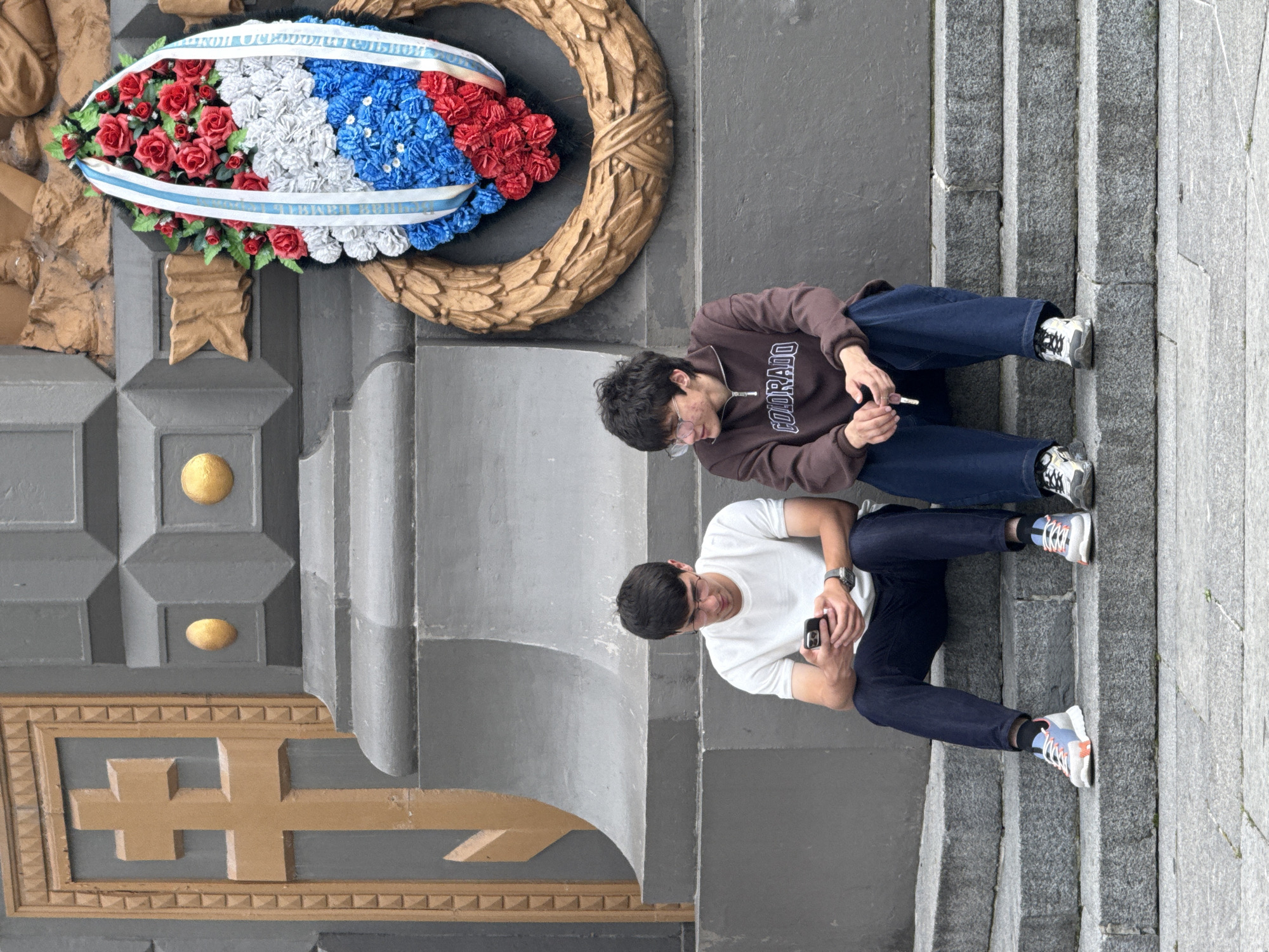 The image shows two young men sitting on stone steps in front of a large, grey, monumental structure. The man on the left, wearing a white t-shirt and dark jeans, is looking at his phone. He has short dark hair and is wearing glasses. The man on the right, wearing a brown sweatshirt with "COLORADO" printed on it and dark jeans, is holding a cigarette. He also has dark hair and is wearing glasses. Behind them, a wreath with red, white, and blue flowers is attached to the structure, suggesting a memorial or a place of remembrance. The structure itself is adorned with a gold cross and other decorative elements. The overall impression is a somber or reflective atmosphere. The architectural style and the presence of the wreath suggest this location could be a war memorial or a significant historical site. Given the context of the wreath and potential architectural style, this scene could be located in Moscow, Russia, possibly near a memorial complex or a significant monument.