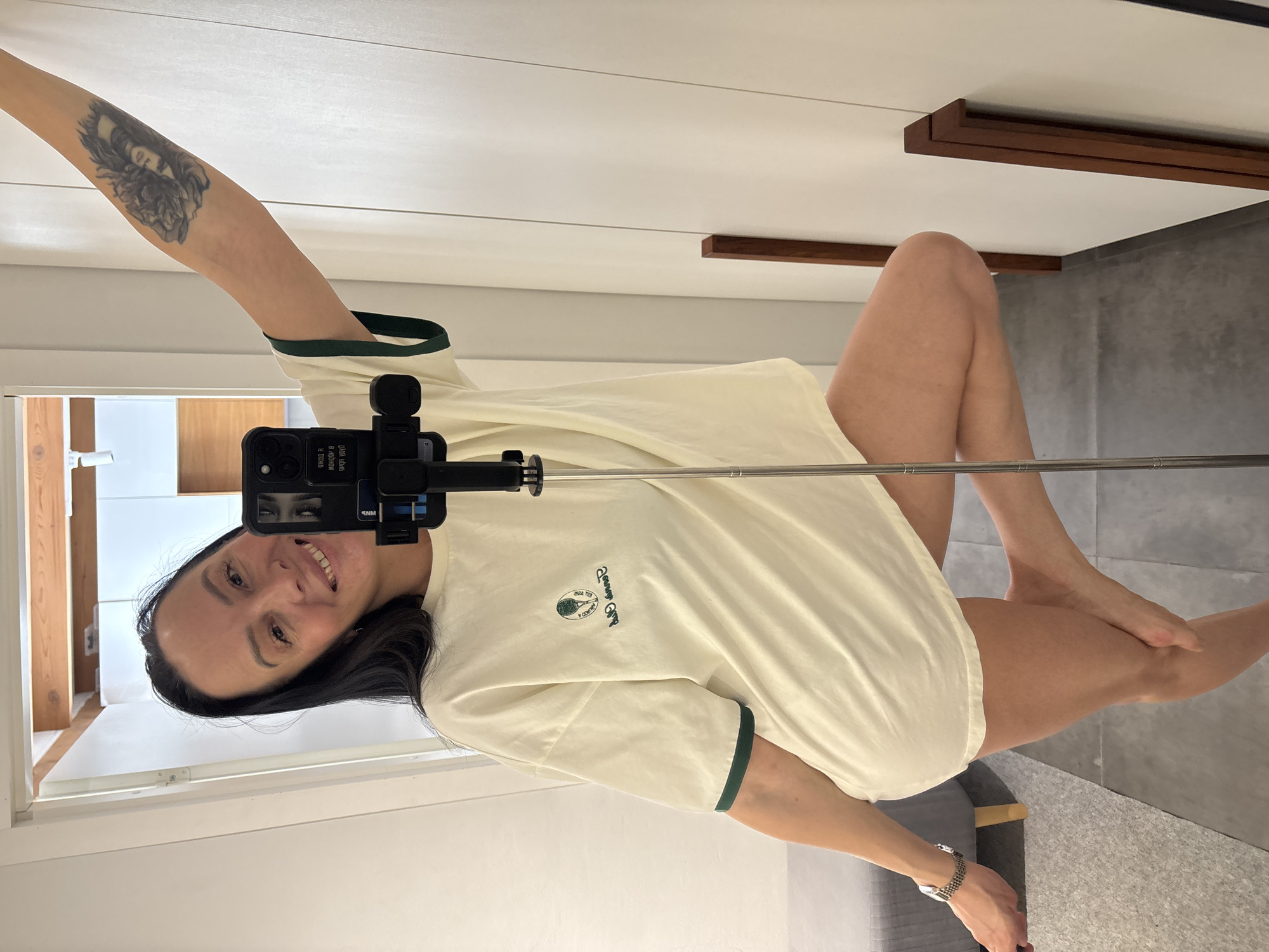 An adult female is depicted taking a mirror selfie indoors in Chaikovsky, Russia. She stands barefoot on a dark grey tiled floor, smiling towards the camera reflection in a full-length mirror. She wears an oversized, light yellow t-shirt with dark green trim on the collar and sleeves, featuring an embroidered "Tennis Club" logo with a tennis racket and ball on the left chest. A tattoo, depicting a woman's face within a floral motif, is visible on her raised left forearm. A metallic watch is present on her right wrist. She balances on her right leg, with her left leg bent and foot raised behind her. She holds a black selfie stick with a smartphone attached, positioned between herself and the mirror. The immediate environment includes light-colored walls, a light grey upholstered bench or sofa to the left, and a white paneled wardrobe with dark brown wooden vertical accents to the right. A reflected background view through an open doorway reveals a room with a white ceiling, exposed wooden beams, and white cabinetry.