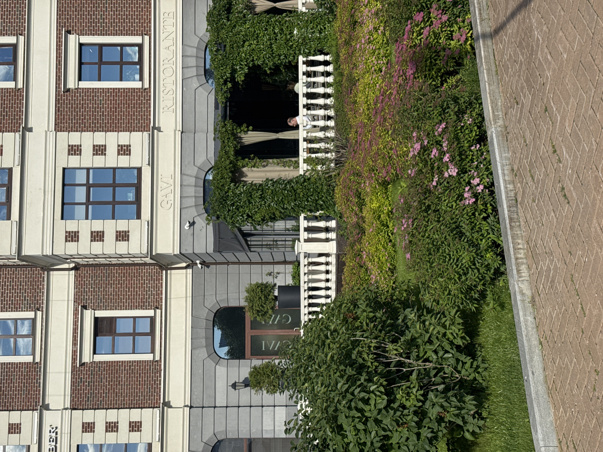 The image depicts the exterior of a multi-story building with a brick facade and stone accents. The ground floor features large arched windows and an entrance area adorned with ivy. Text on the building reads "GAVI" and "RISTORANTE". A white balustrade separates the building's patio from a lush garden filled with various green shrubs and pink flowers. In the background, a person is seated at a table on the patio. The building is located in Yekaterinburg, Russia, specifically at the intersection of Malysheva Street and Belinsky Street, housing the restaurant "Gavi" and "Ristorante".
