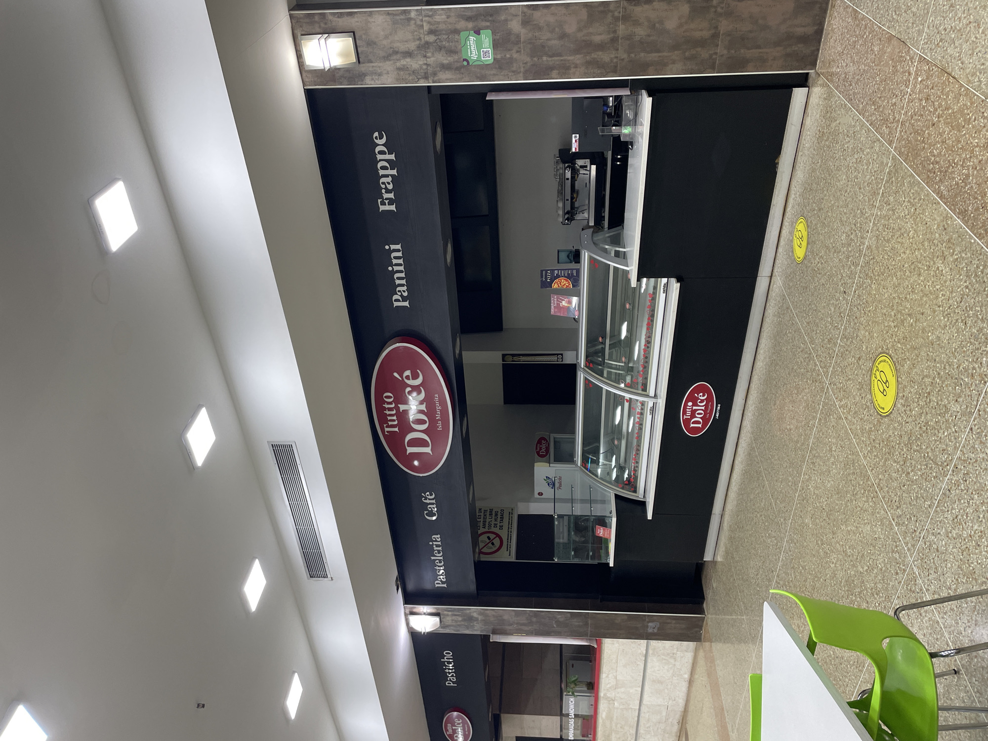 The image shows the exterior of a food establishment in El Valle del Espíritu Santo, Venezuela. The establishment, named "Tutto Dolce," appears to be a cafe and pastry shop. It features a black facade with illuminated signs indicating offerings such as "Pasteleria Café," "Panini," and "Frappe." A prominent red oval logo with "Tutto Dolce" and "Isla Margarita" is visible. In front of the counter, there are display cases filled with food items, likely pastries or desserts. Behind the counter, equipment such as a coffee machine and a cash register are present. The floor is tiled with a speckled pattern, and yellow social distancing markers are visible on the ground. To the left, there are tables and green chairs, suggesting a seating area. The overall environment is indoors, likely a shopping mall or commercial complex, with overhead lighting fixtures.